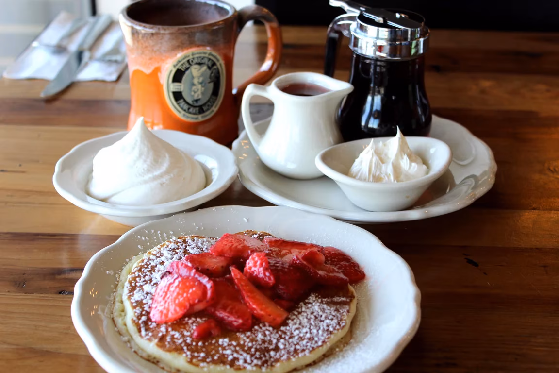 Pancakes with strawberries, powdered sugar, whipped cream, syrup, and coffee mug.