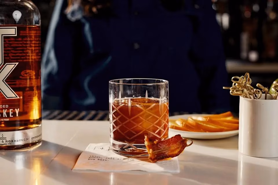 Whiskey bottle, cocktail with bacon, and orange slices at a bar.