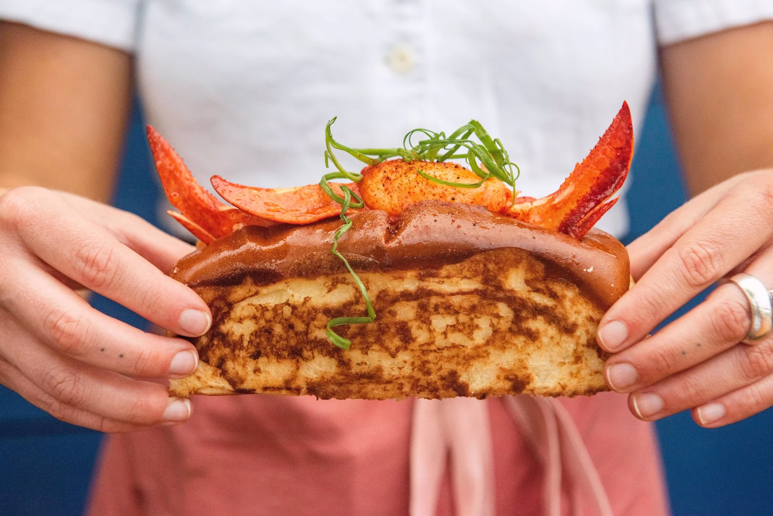 Hands hold a lobster roll on a toasted bun with green garnish.