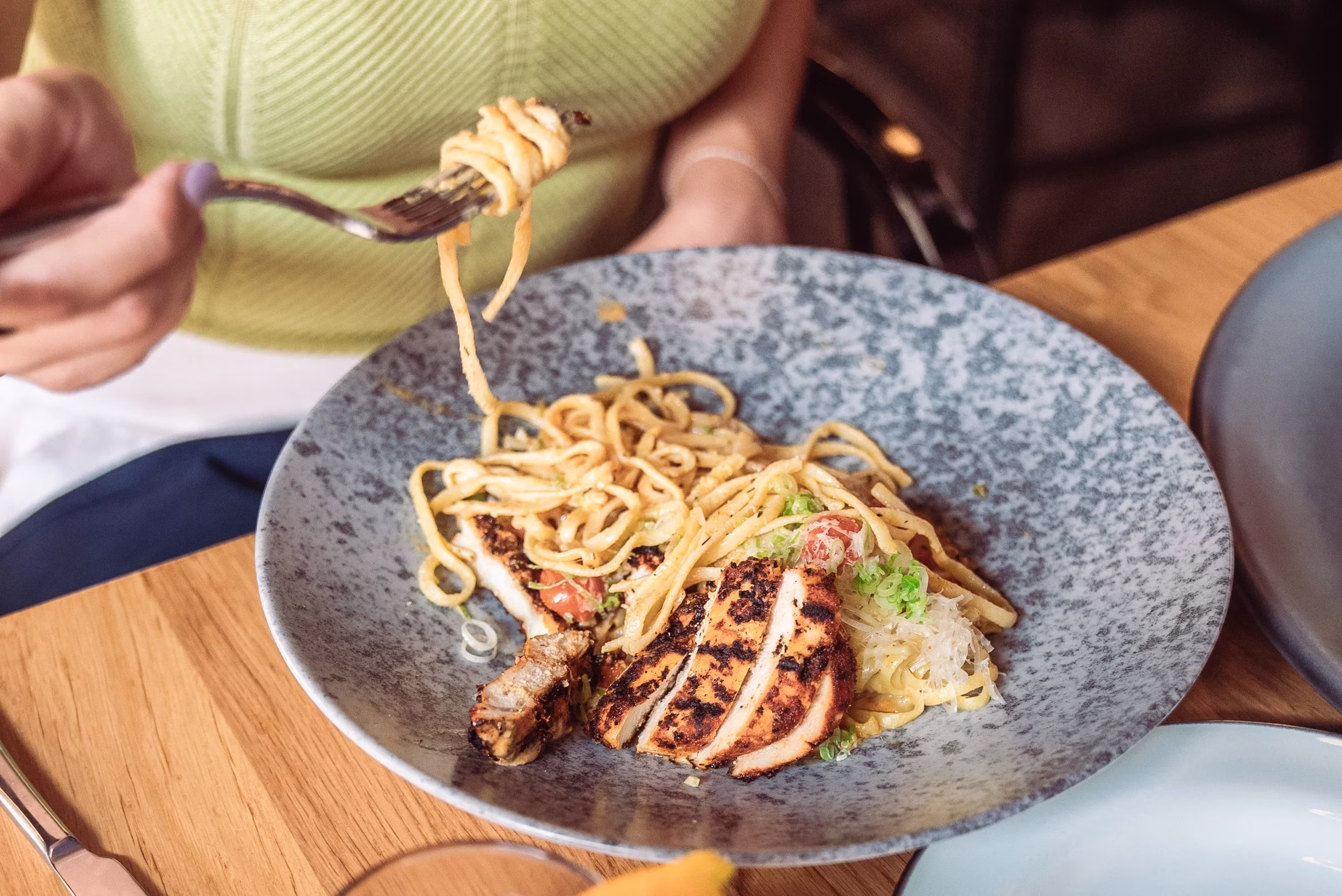 Hand with fork lifting noodles from a plate of grilled chicken.