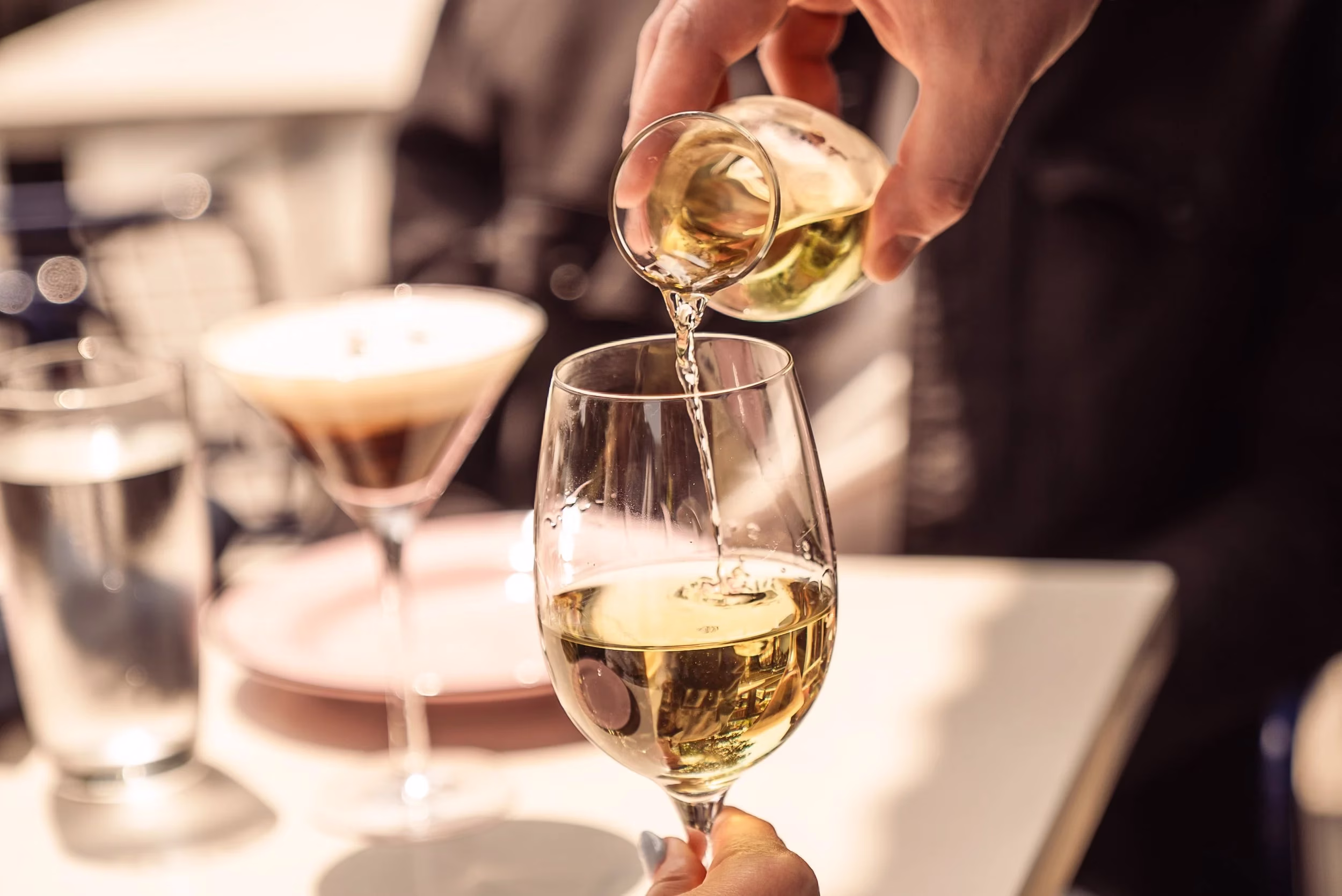 Hand pouring light-colored liquid from carafe into a wine glass.