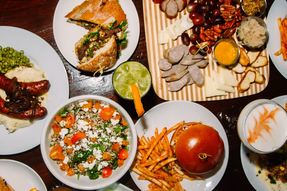Table with multiple plates of sausages, charcuterie, salad, burger, and drinks.