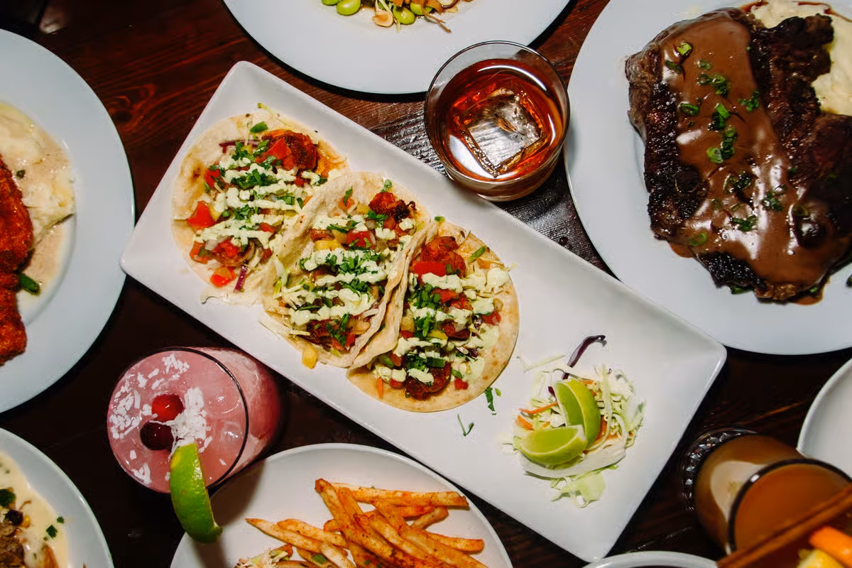 Table laden with tacos, steak, mashed potatoes, fries, and cocktails.