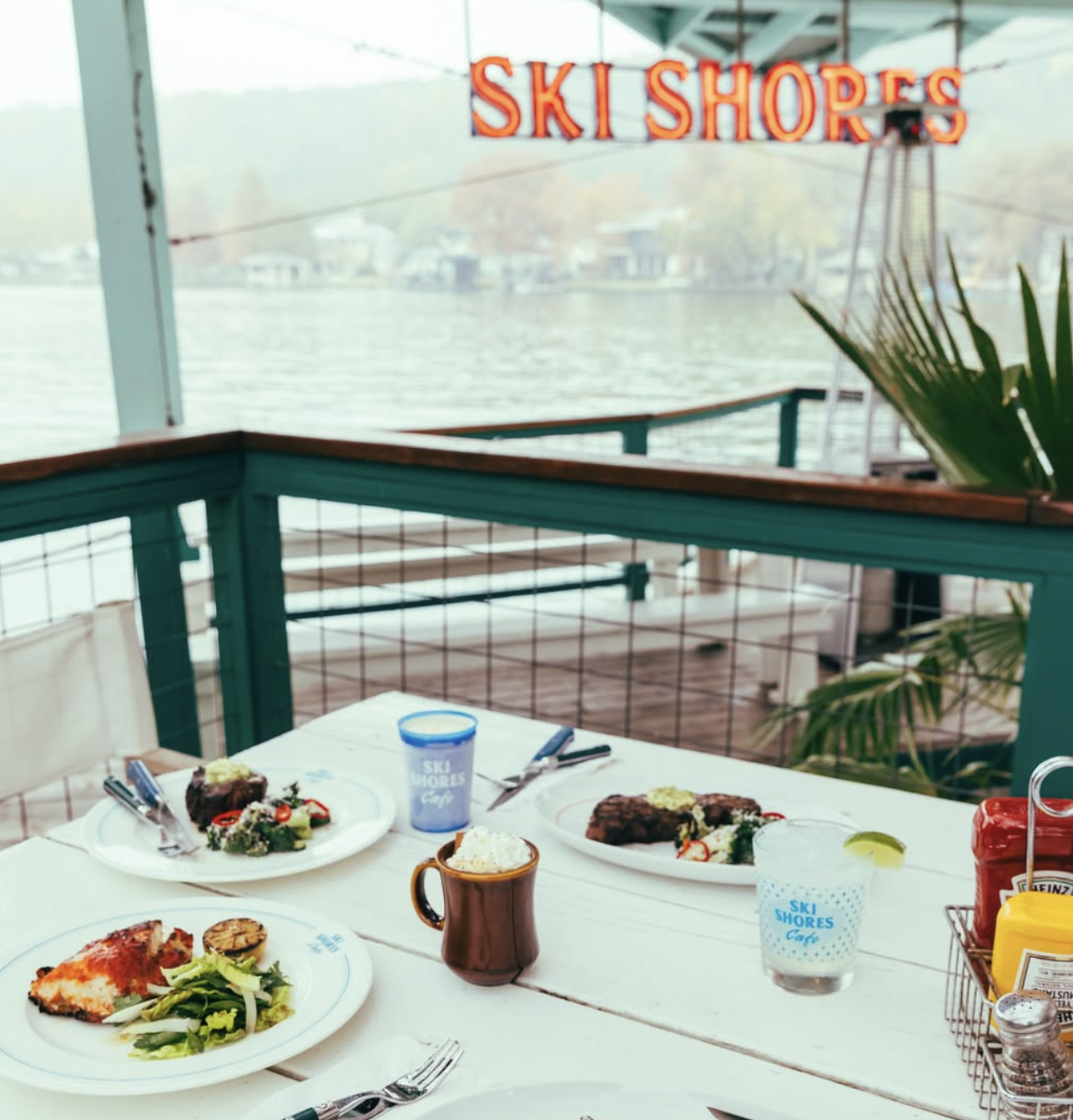 Meals and drinks on a table at Ski Shores Cafe overlooking a lake.