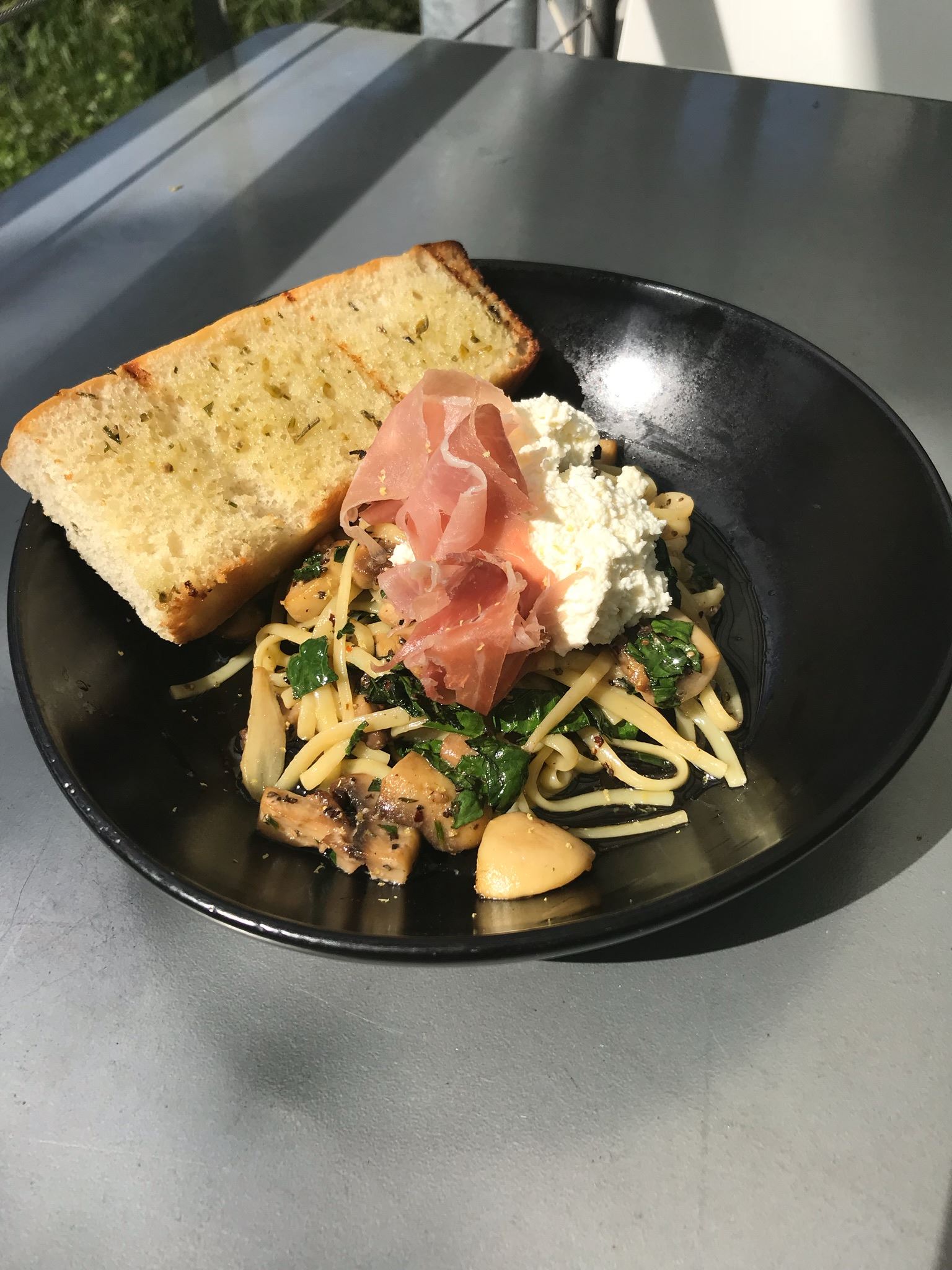 Black plate of pasta with mushrooms, spinach, prosciutto, ricotta, and garlic bread.