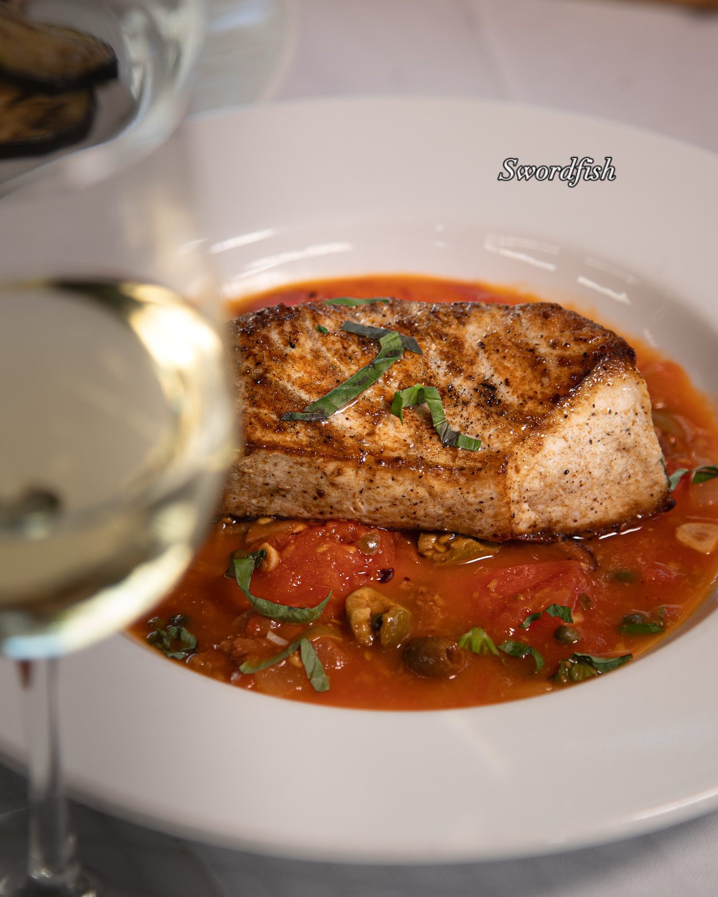 Seared swordfish steak in tomato sauce, basil, with blurred wine glass.