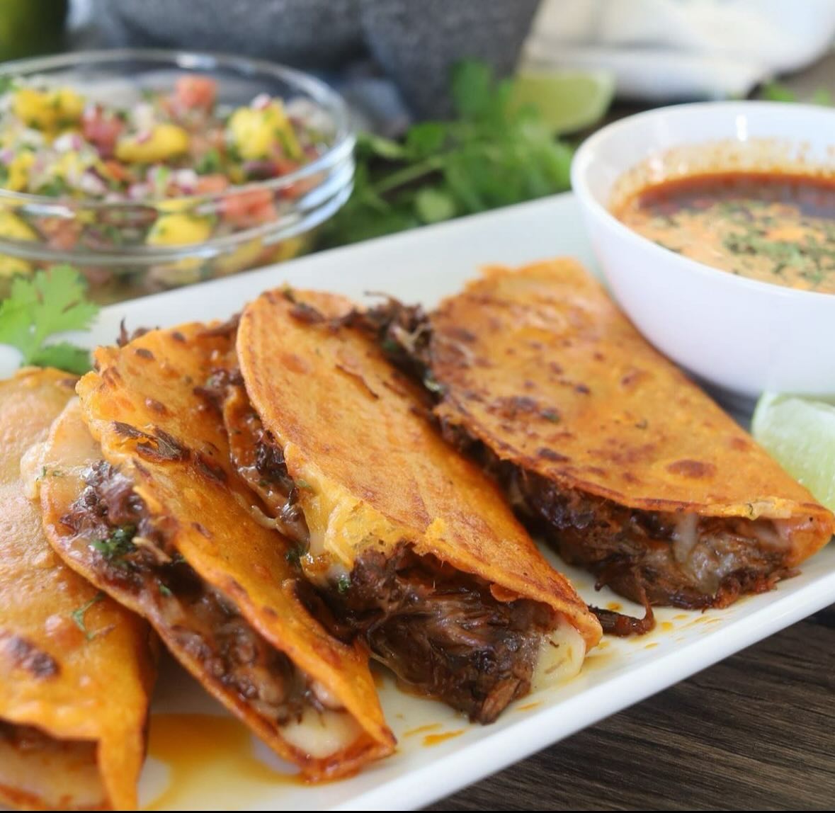 Crispy birria tacos, dipping sauce, and mango salsa on a table.