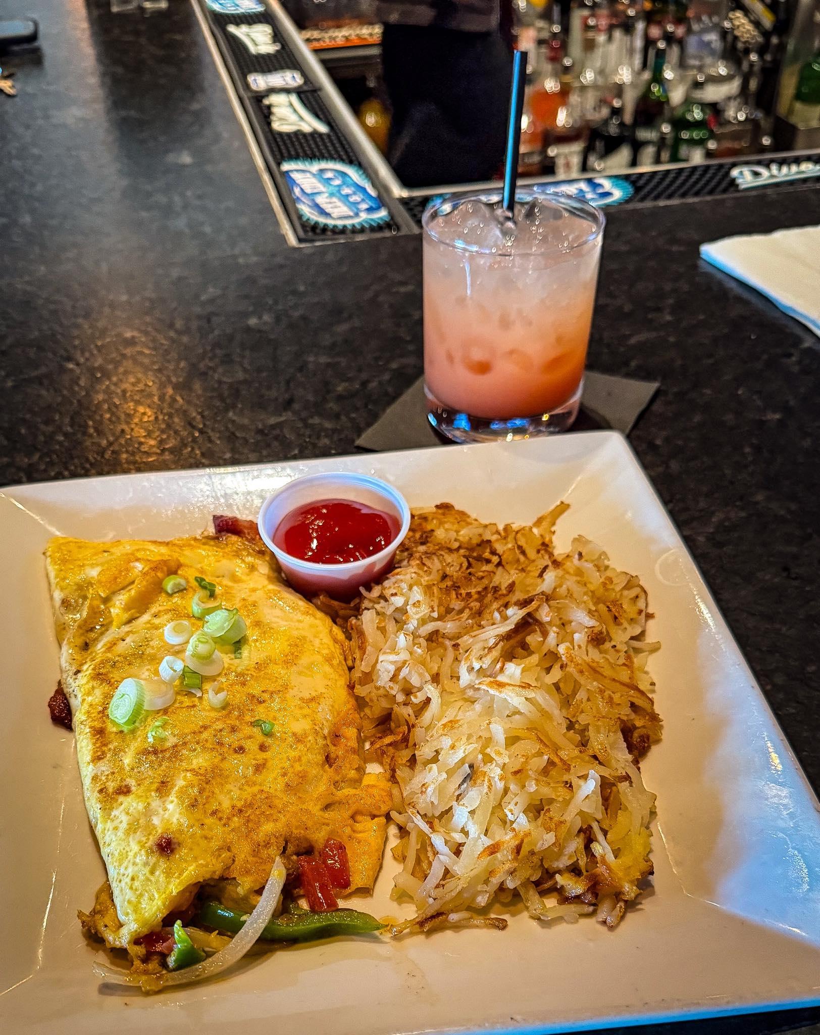 Omelet, hash browns, ketchup, and cocktail on a bar counter.