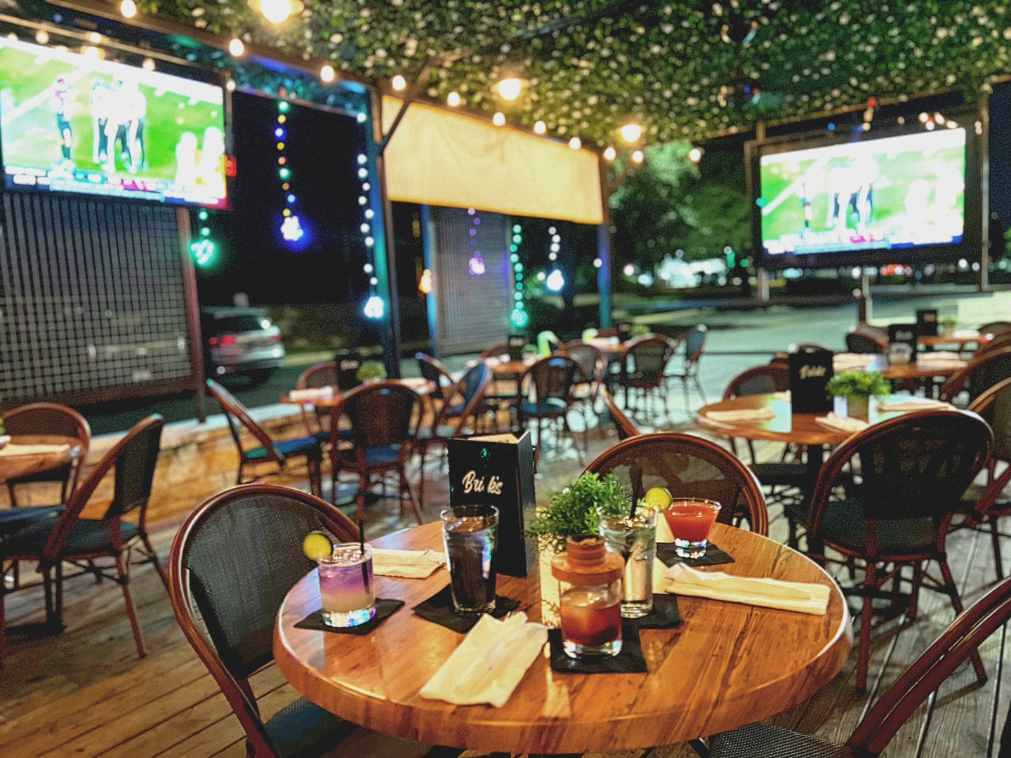 Nighttime outdoor patio with tables, drinks, string lights, and sports TVs.