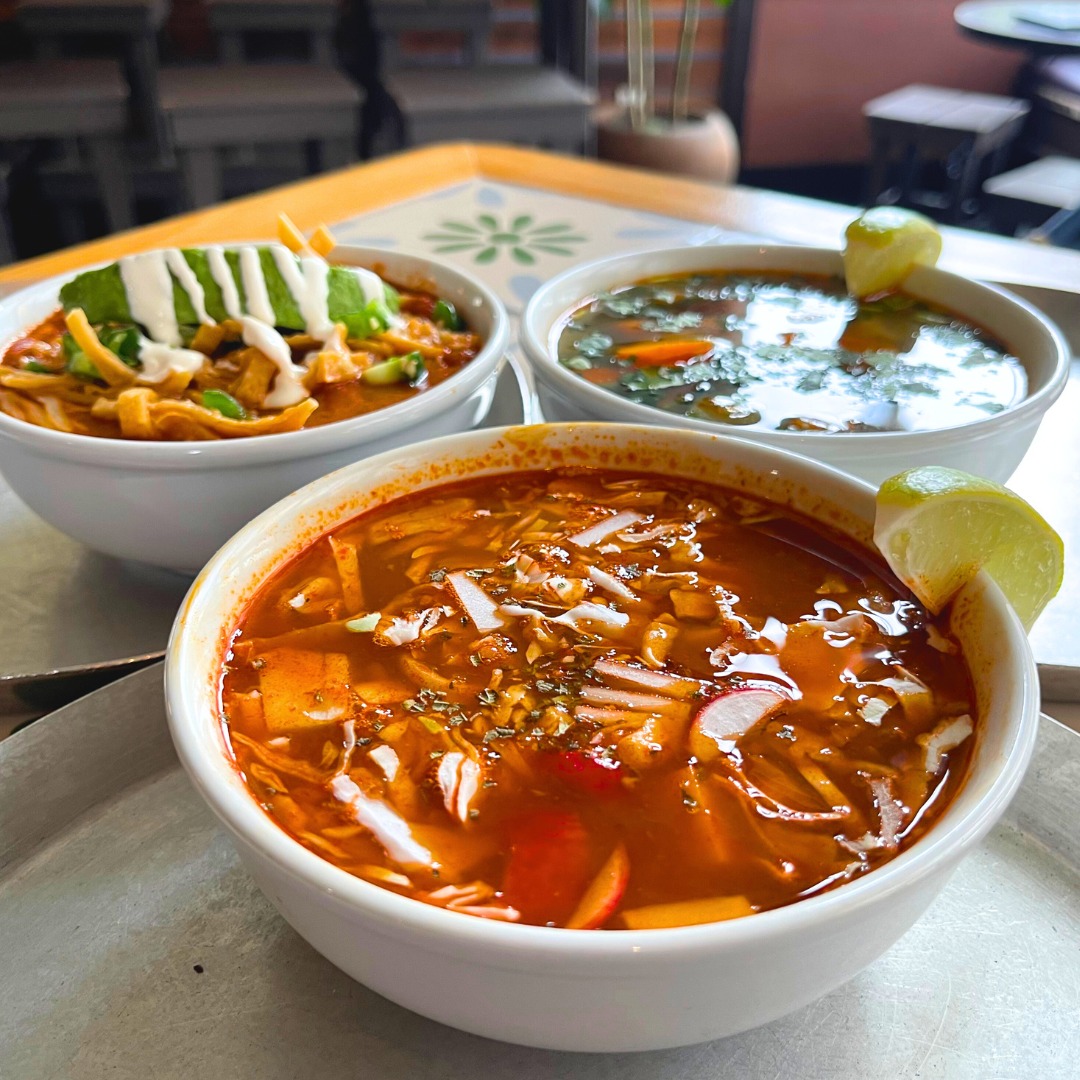 Three bowls of colorful soups, garnished with avocado, lime, and radish.