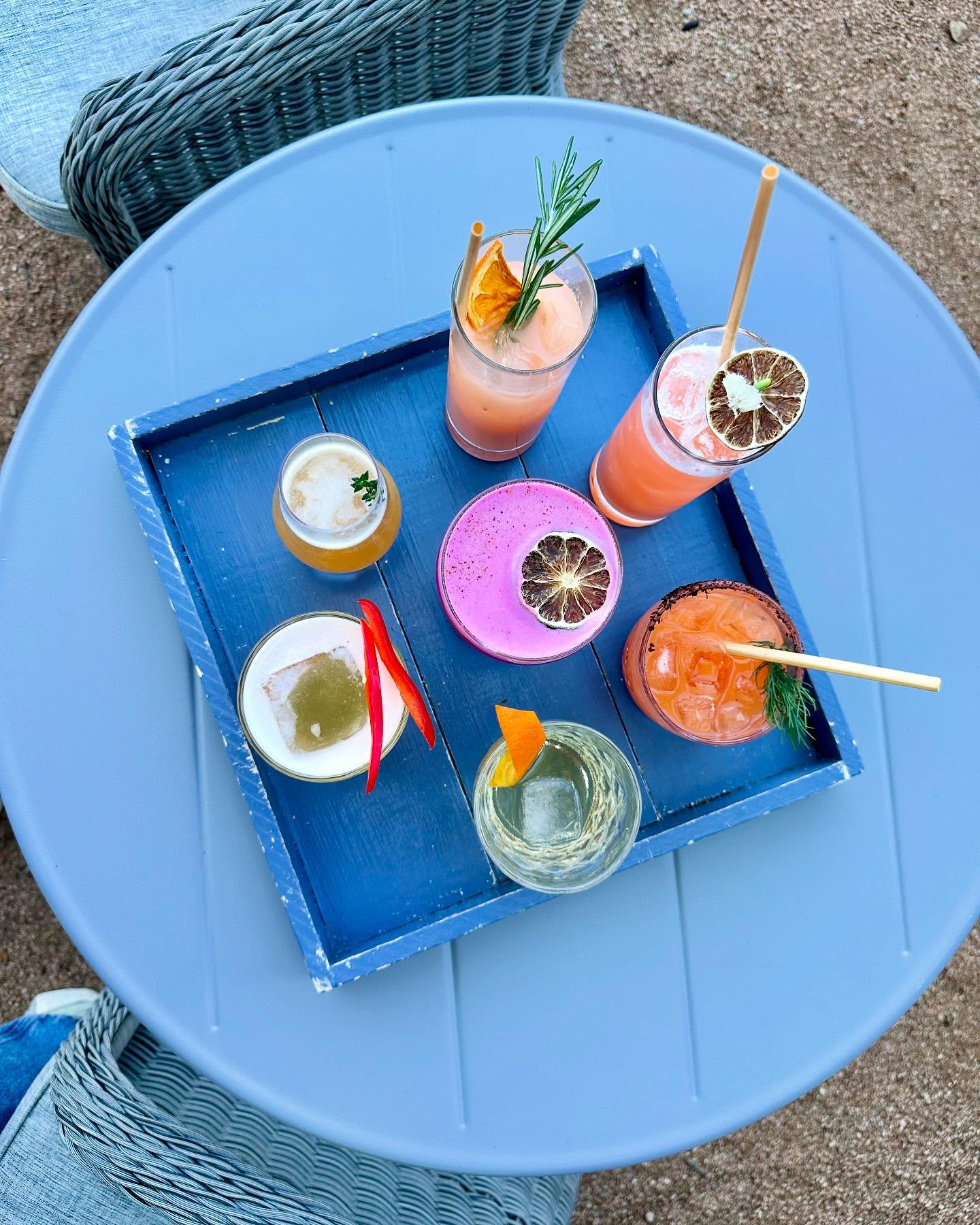 Six colorful cocktails on a blue tray on a light blue table.