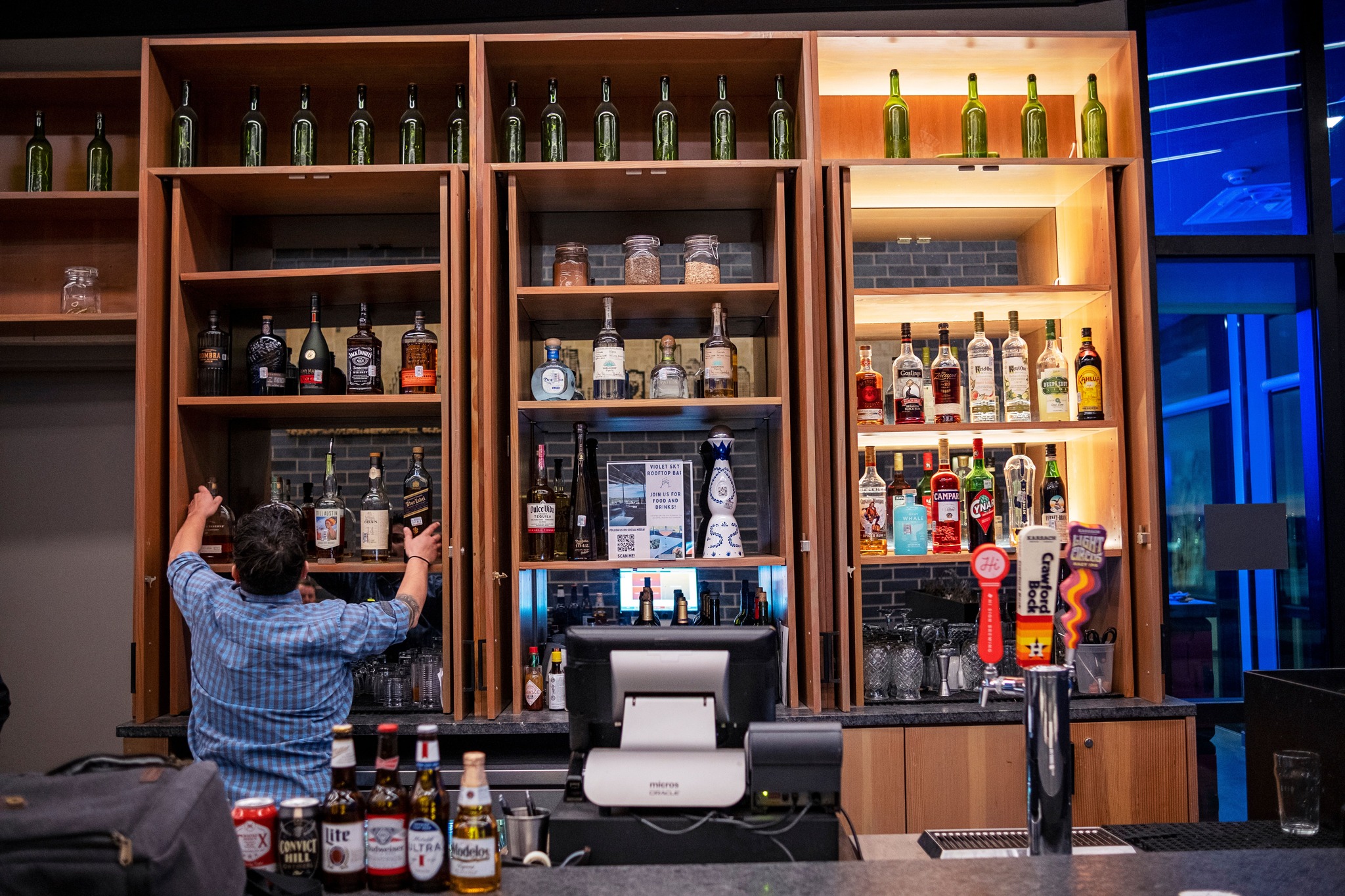 Bartender organizing liquor bottles on illuminated shelves behind a bar.