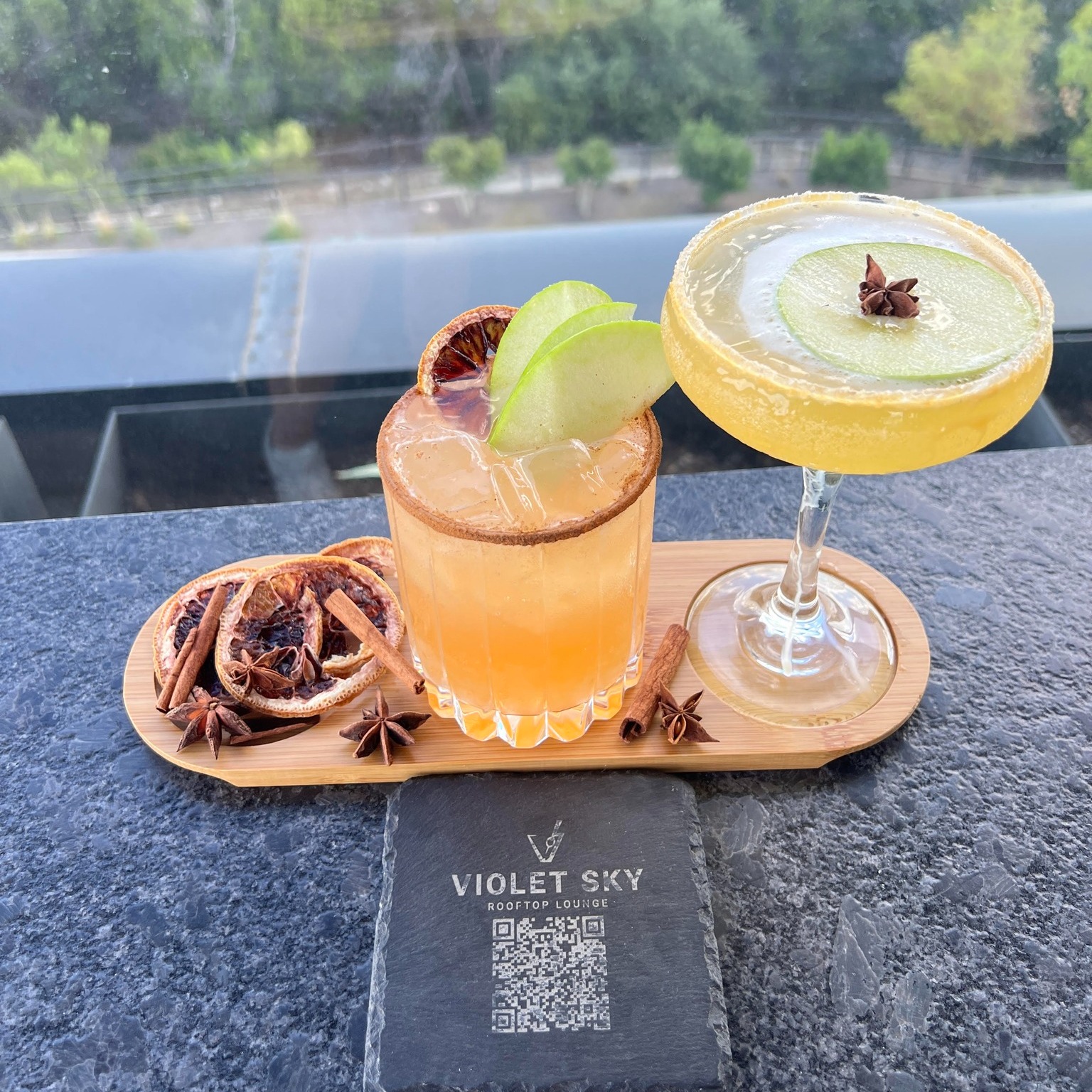 Two cocktails garnished with fruit, spices, and a Violet Sky coaster.