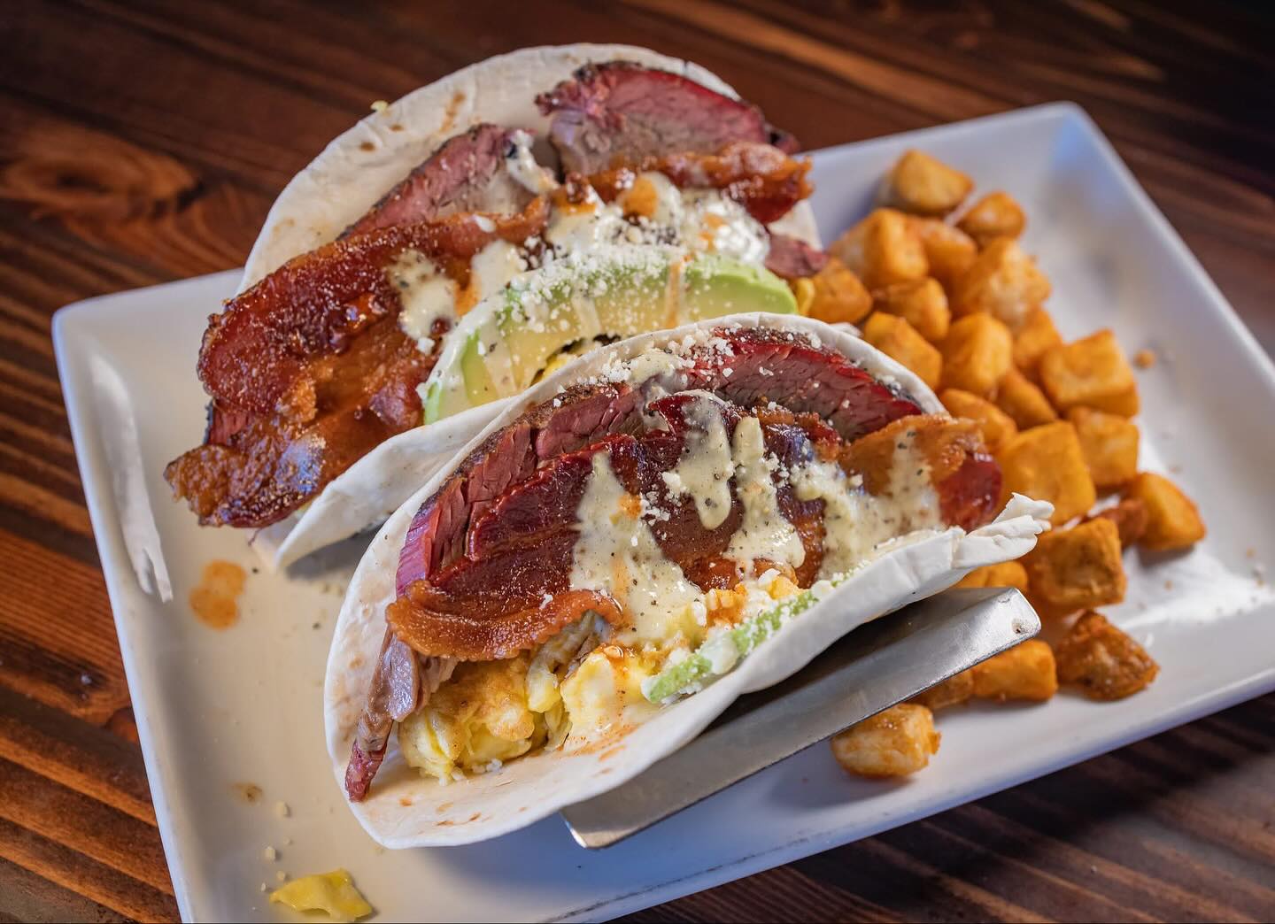 Two breakfast tacos with steak, bacon, avocado, and fried potatoes.