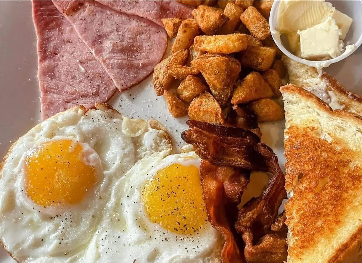 Breakfast plate with eggs, ham, bacon, home fries, toast, and butter.