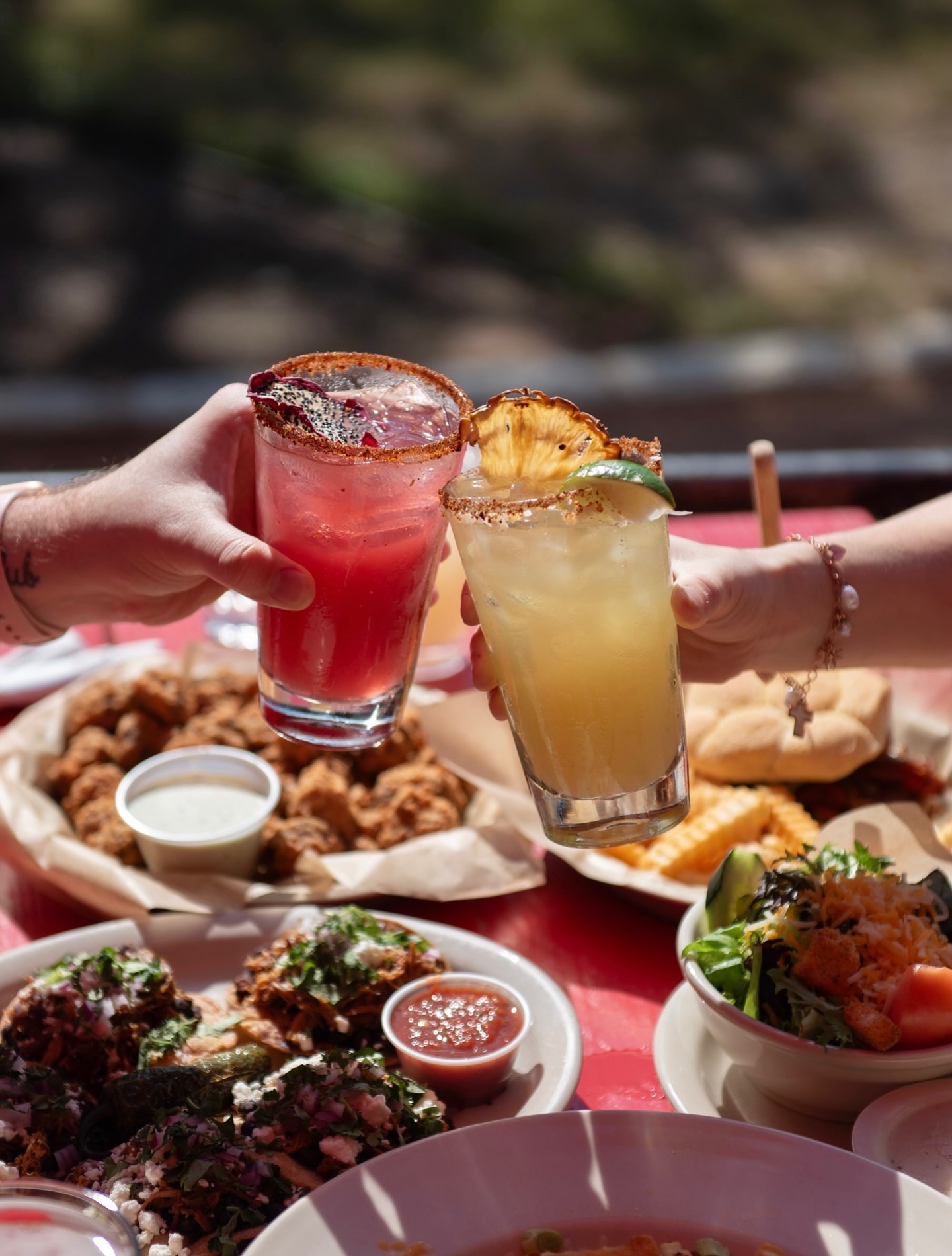 Hands toasting pink and yellow cocktails with many dishes of food.
