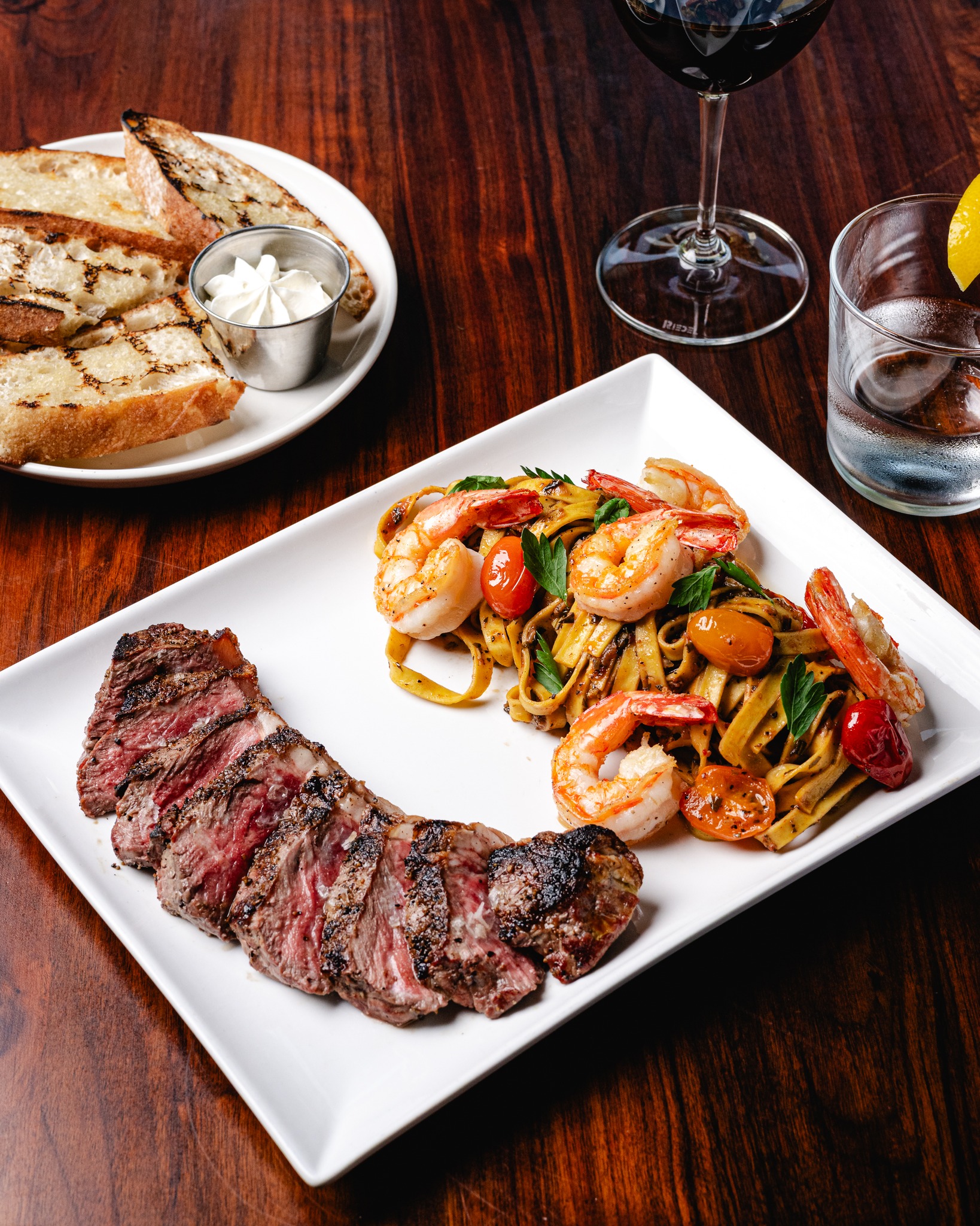 Sliced steak with shrimp pasta, bread, and drinks on a wooden table.