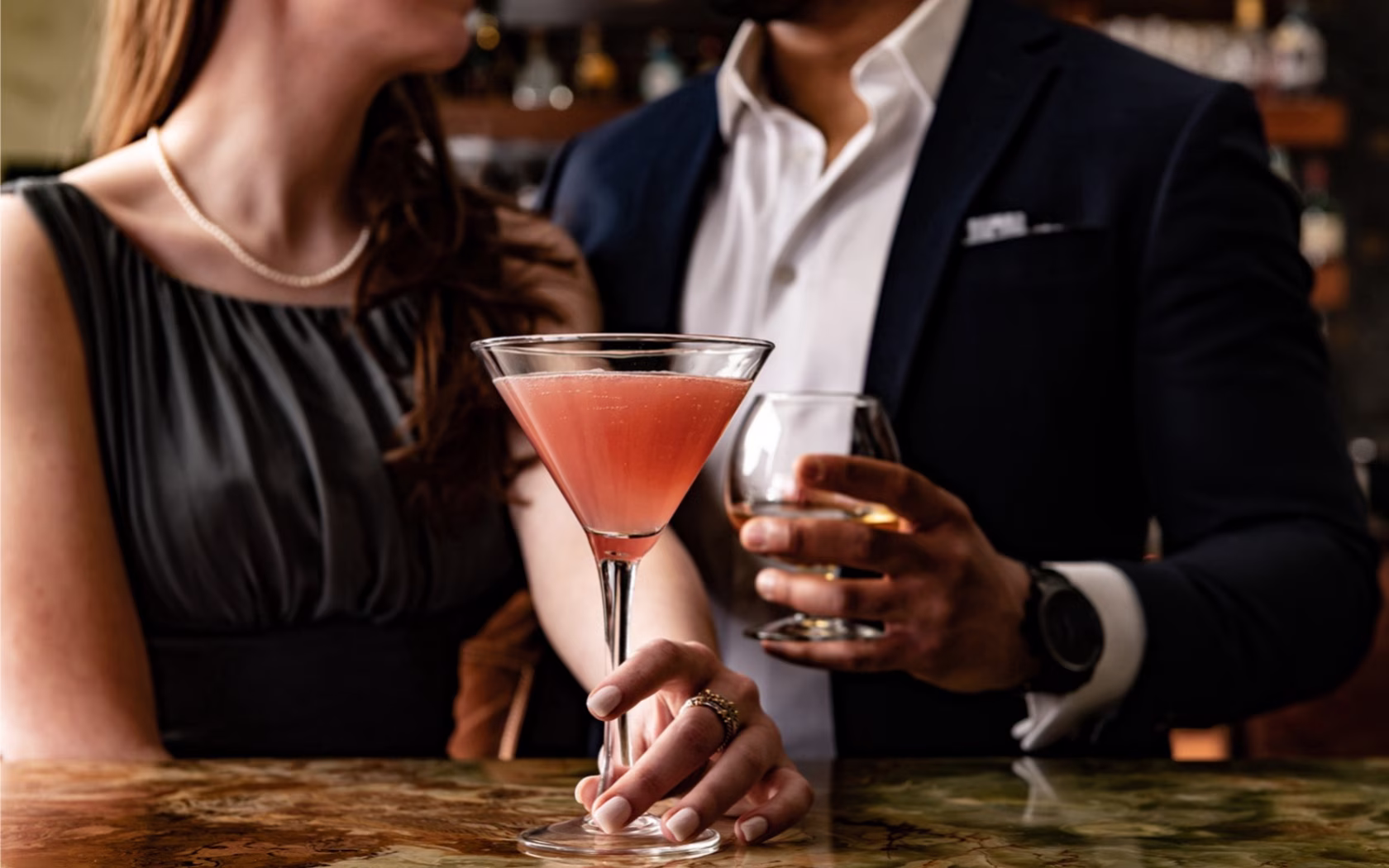 Woman and man holding a pink drink and an amber drink at a bar.