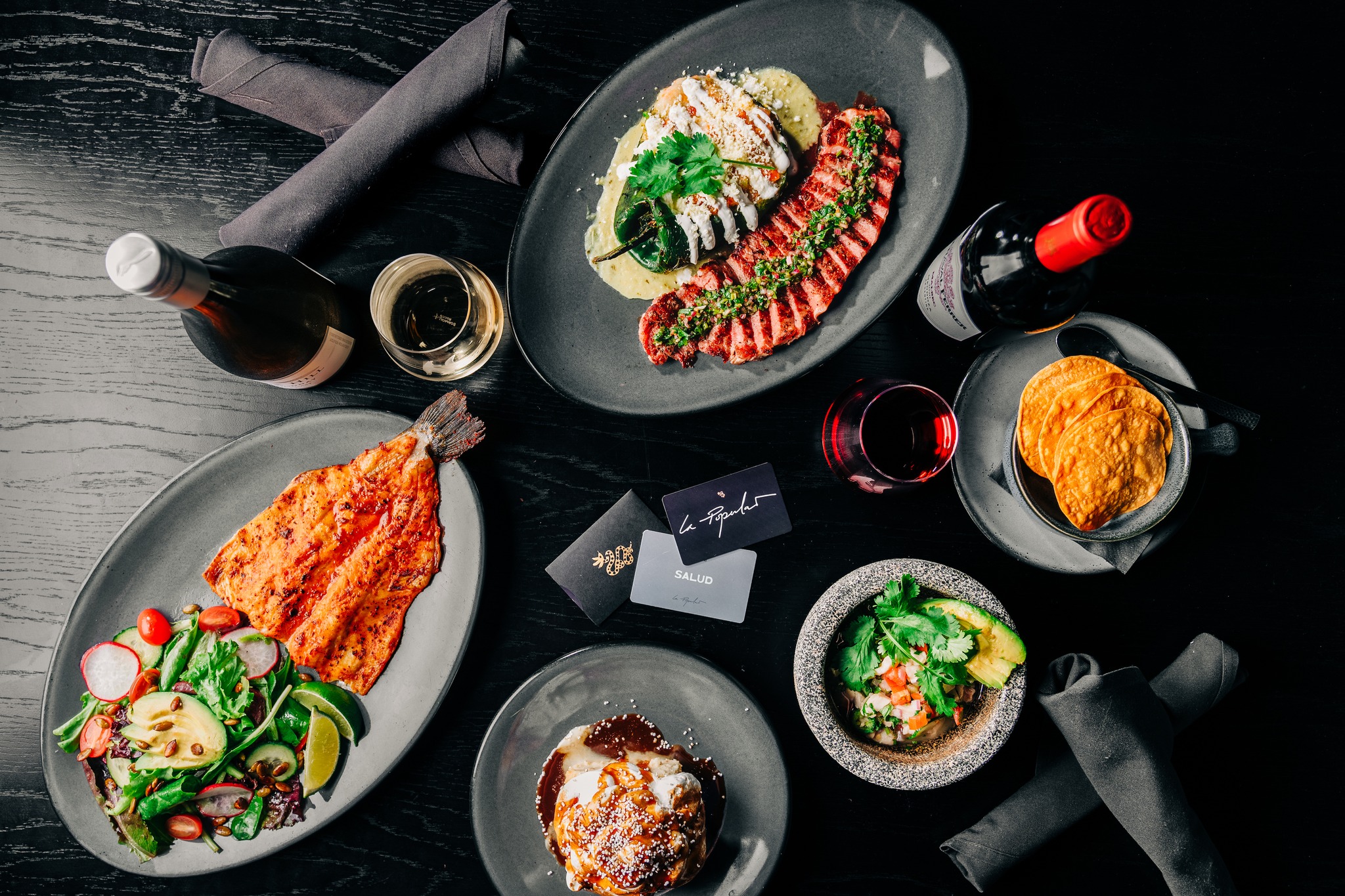 Multiple plates of food, wine, and gift cards on a dark table.