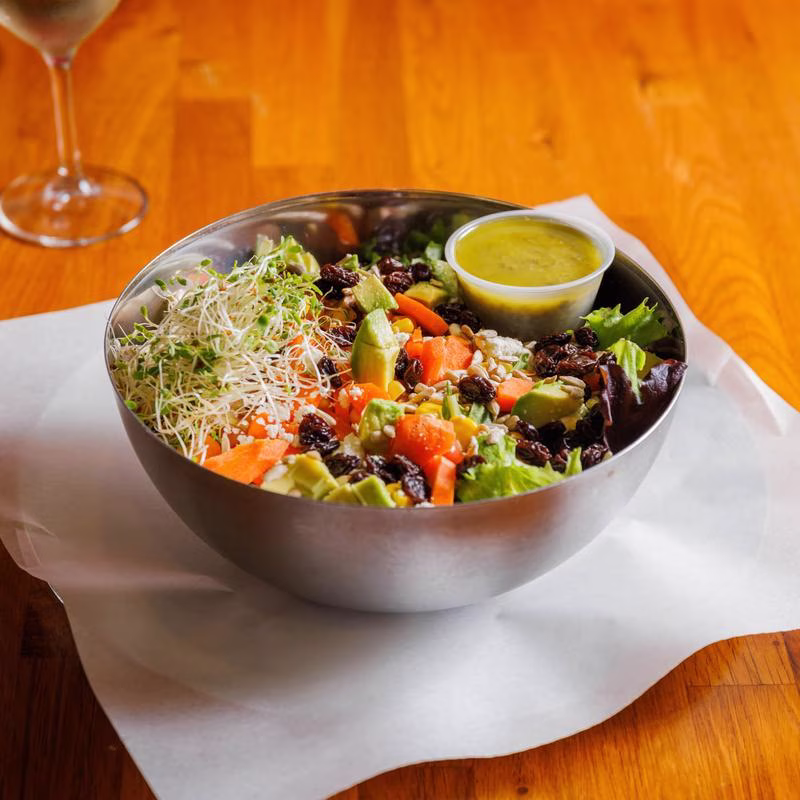 A metal bowl of salad with sprouts, avocado, raisins, and dressing.