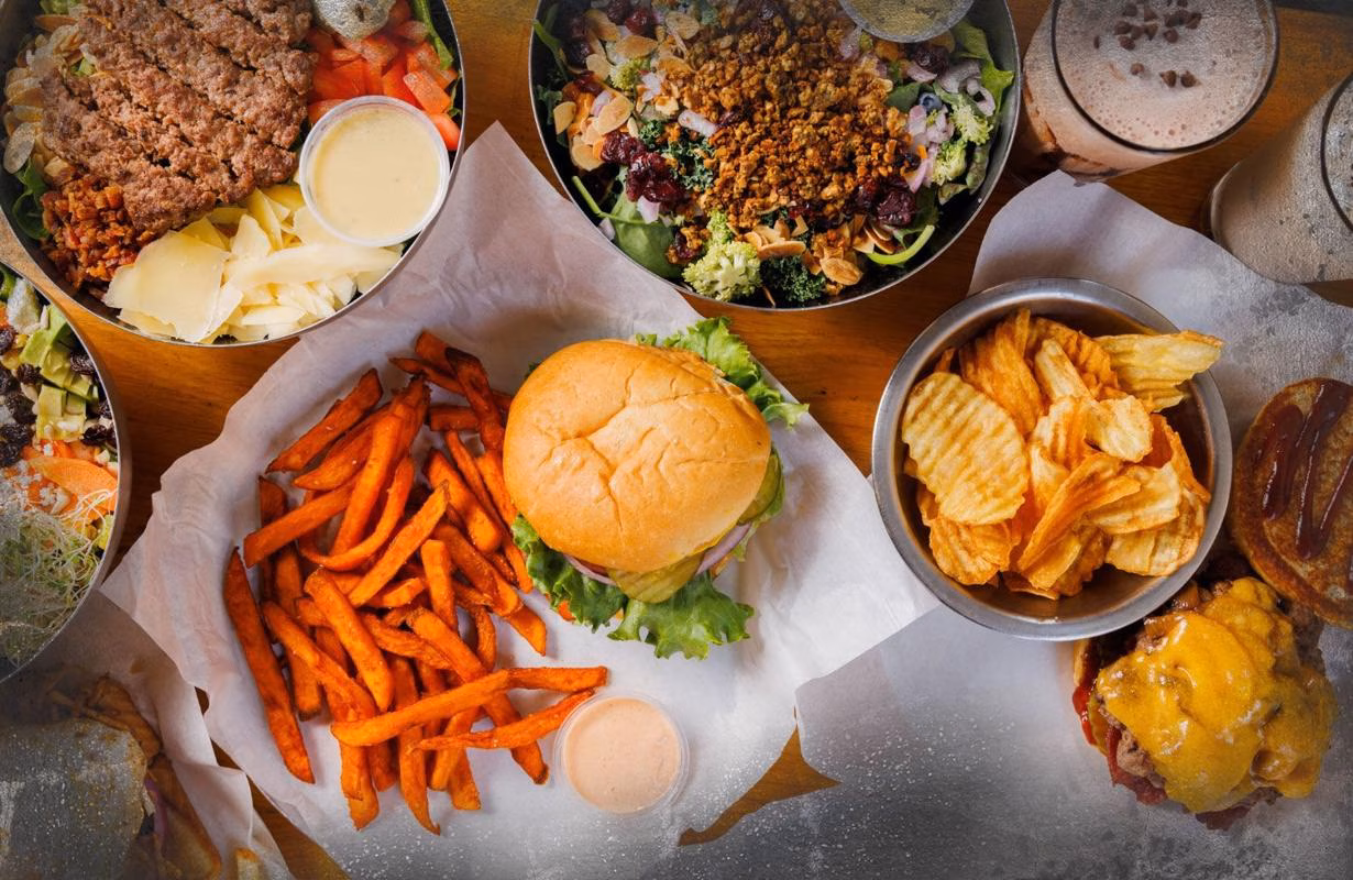 Tabletop full of burgers, salads, sweet potato fries, chips, and drinks.