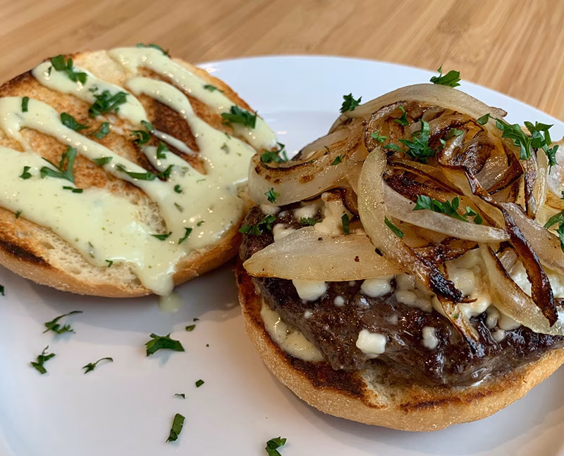 Open-faced burger with patty, crumbled cheese, caramelized onions, herbed sauce.