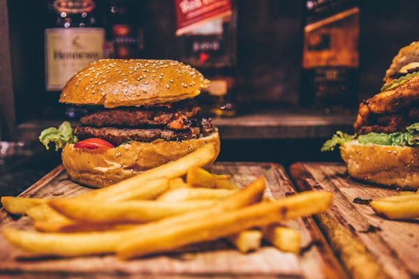 Double burger and fries on wooden board, with blurry alcohol bottles.