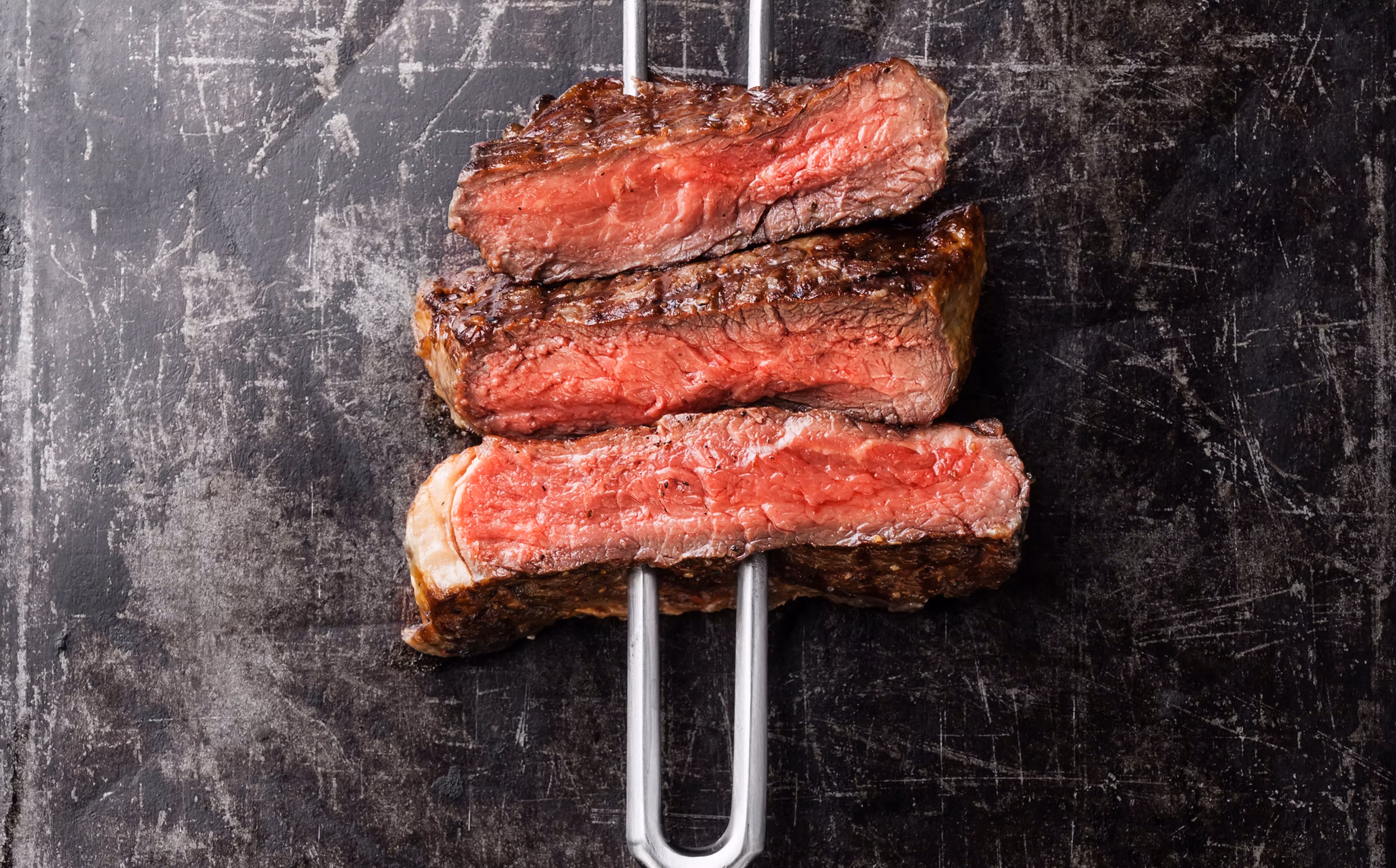 Three medium-rare steak slices skewered on metal rods on dark surface.