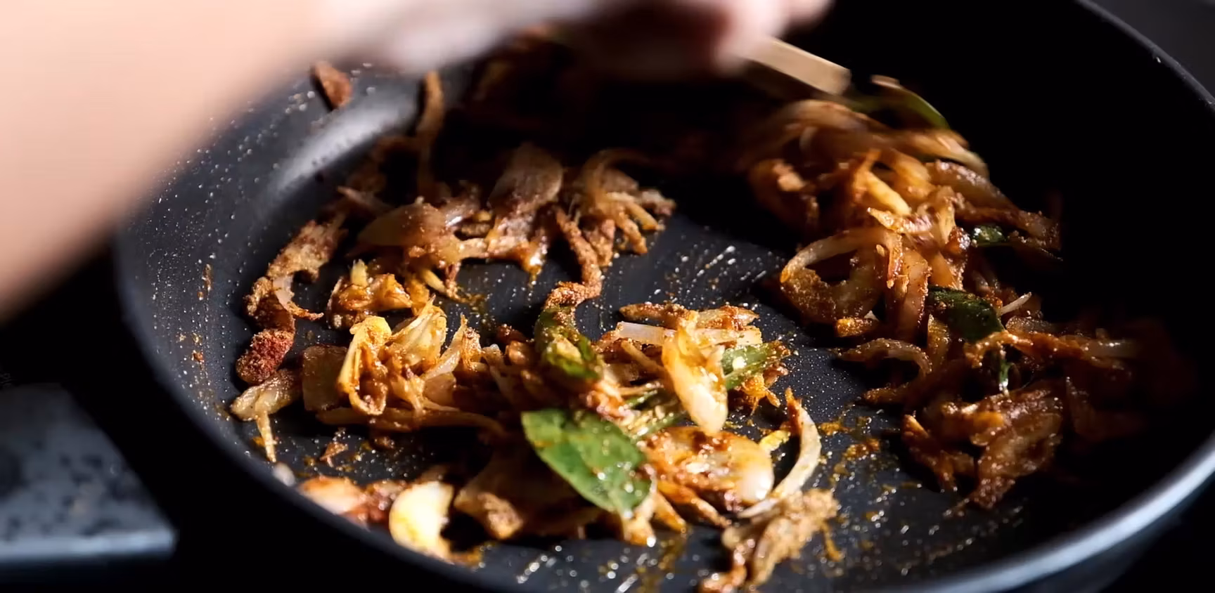 Sliced onions, spices, and green leaves cooking in a frying pan.