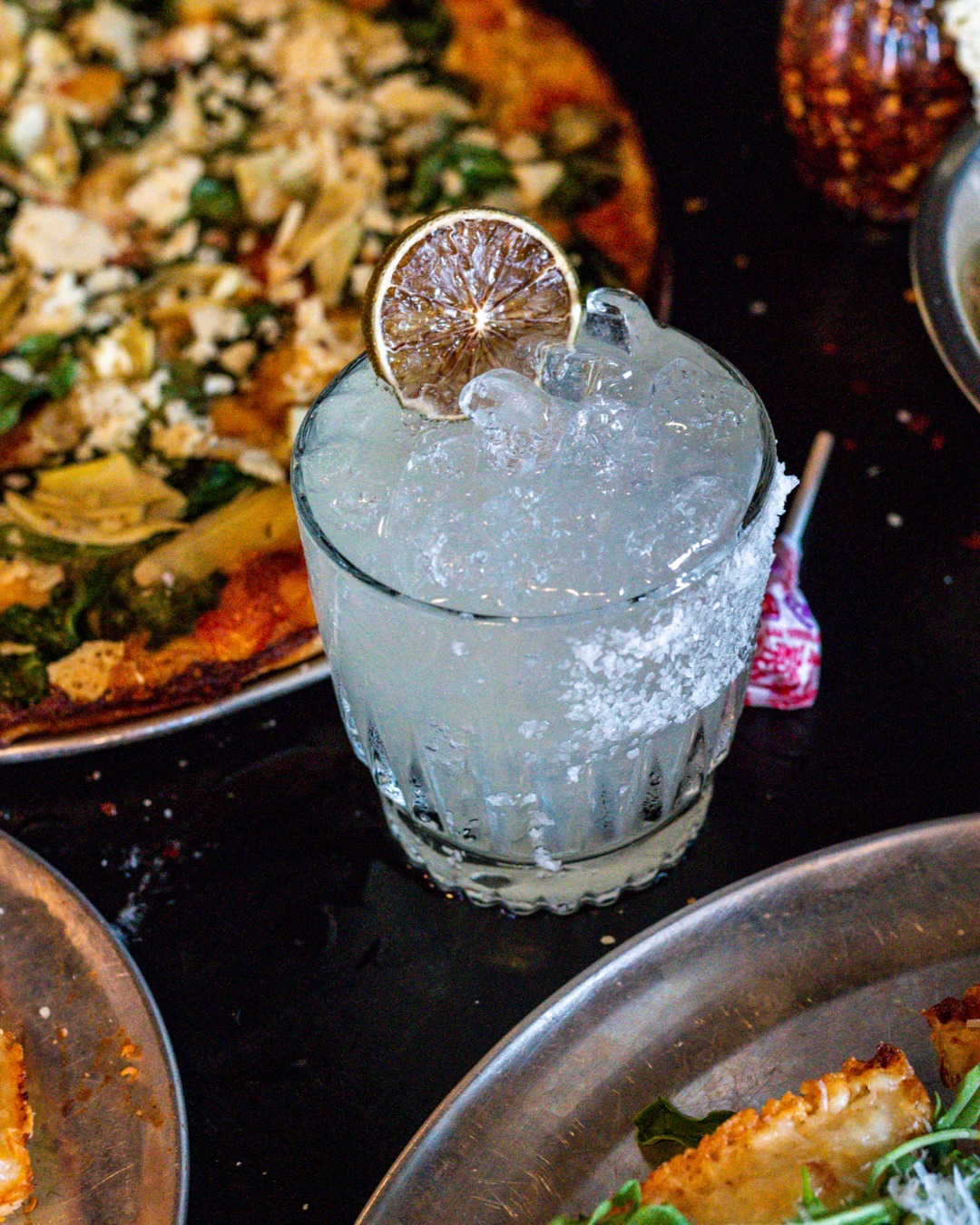 Iced beverage with a dried lime, salted rim, and pizza background.