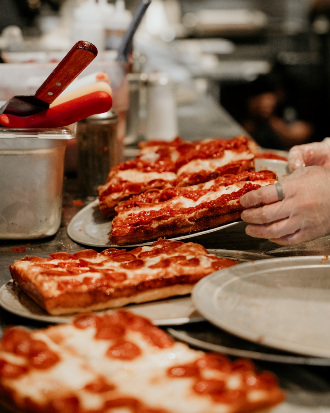 Gloved hand serves thick-crust rectangular pizza slices in kitchen.