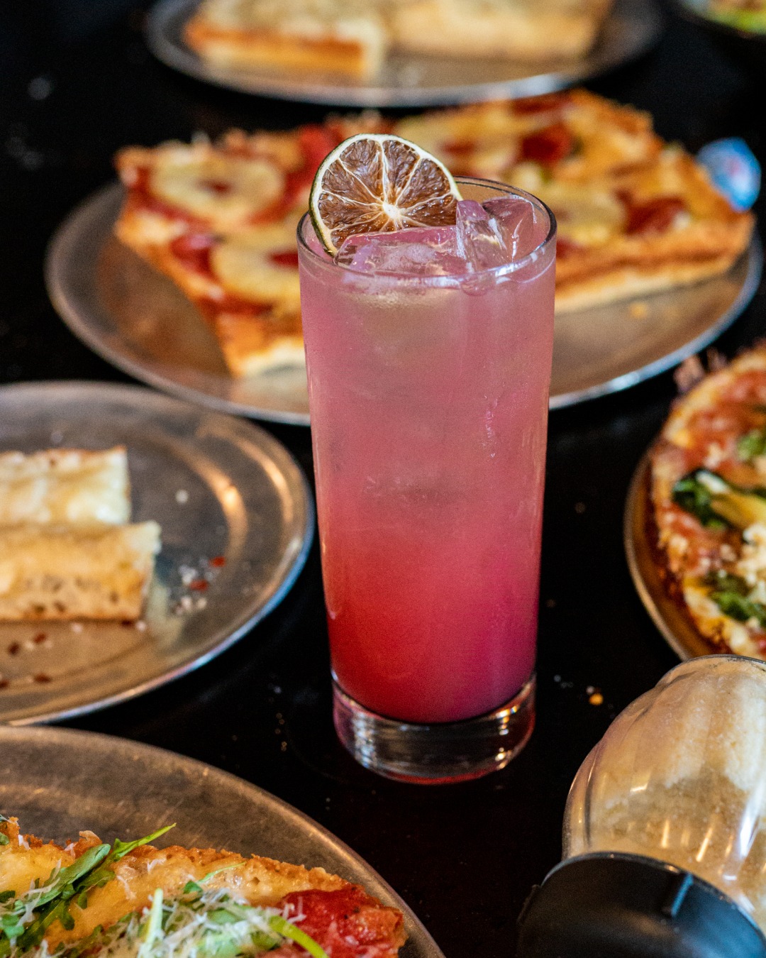 Pink drink with lime, ice, and various pizzas on dark table.