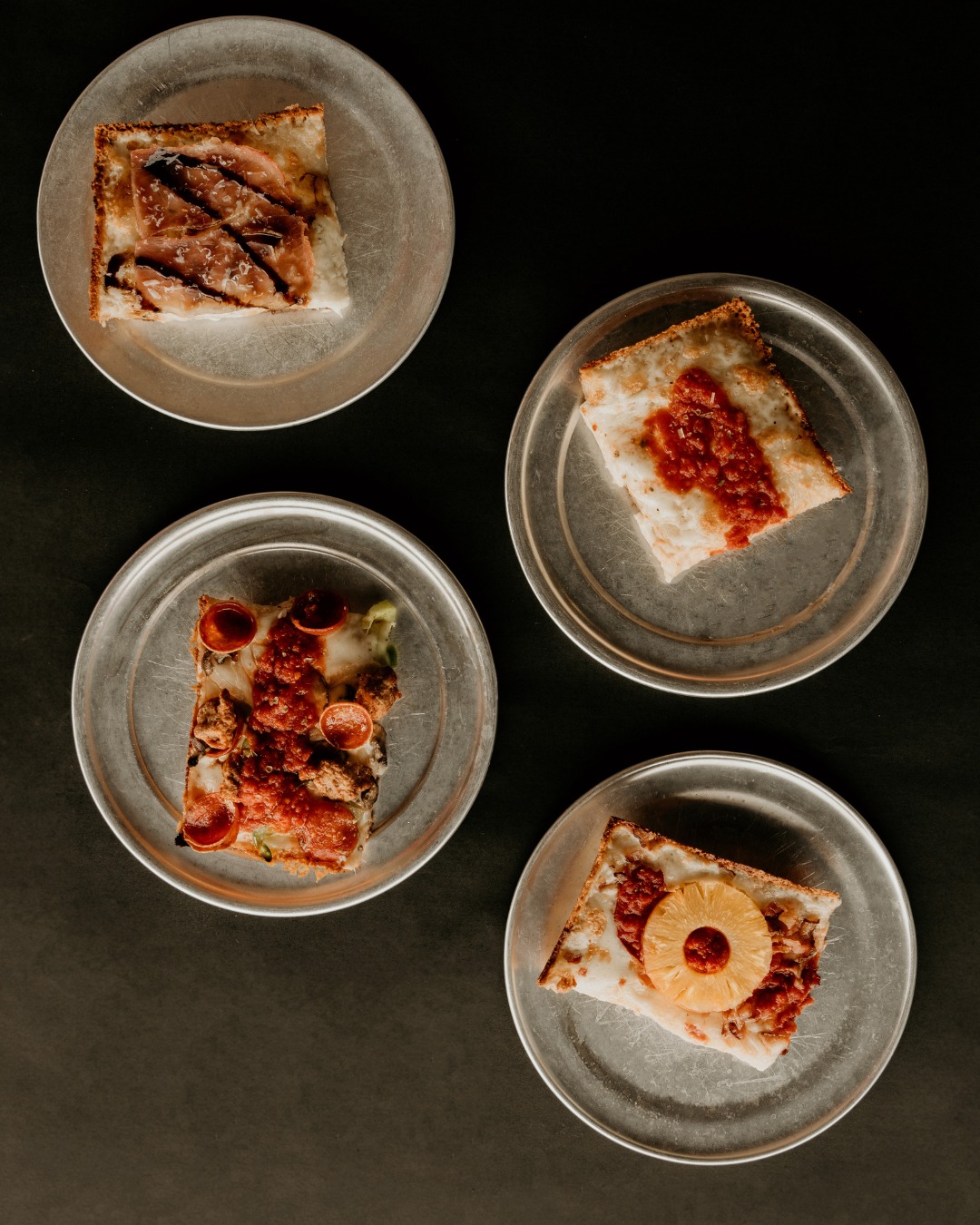 Four different square pizza slices on individual metal plates.