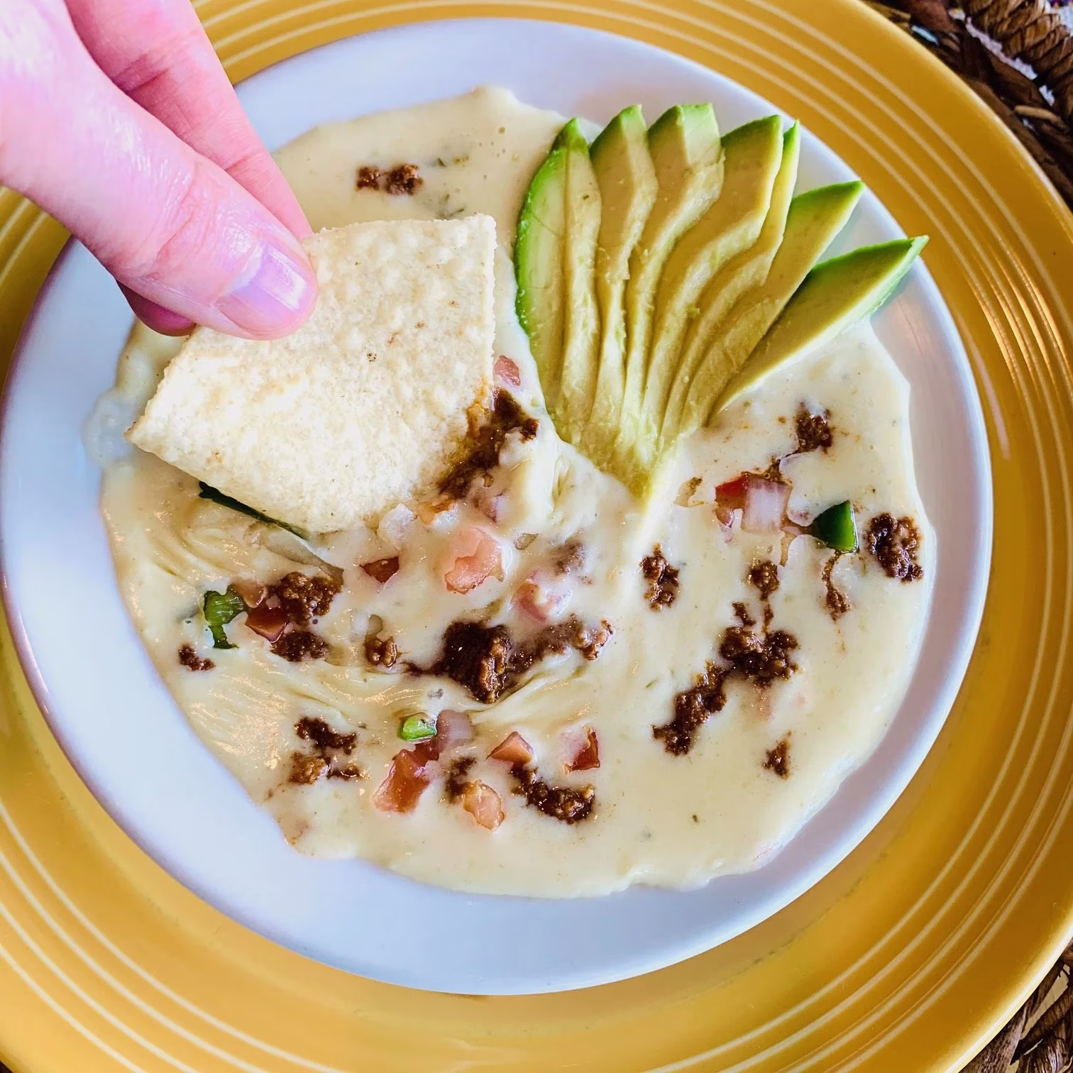 Hand dipping chip into queso with avocado, dark crumbles, and vegetables.