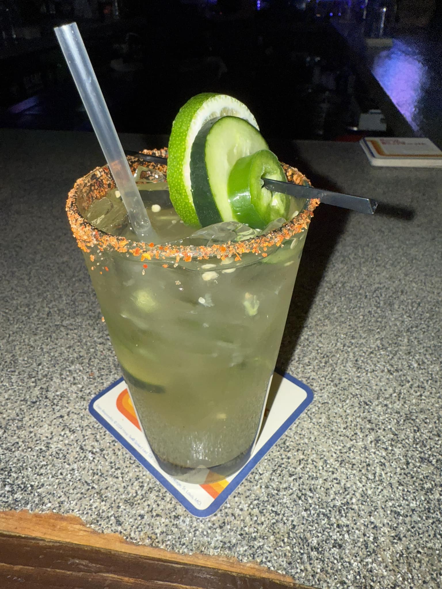 Green drink with spiced rim, lime, cucumber, and jalapeño on bar.