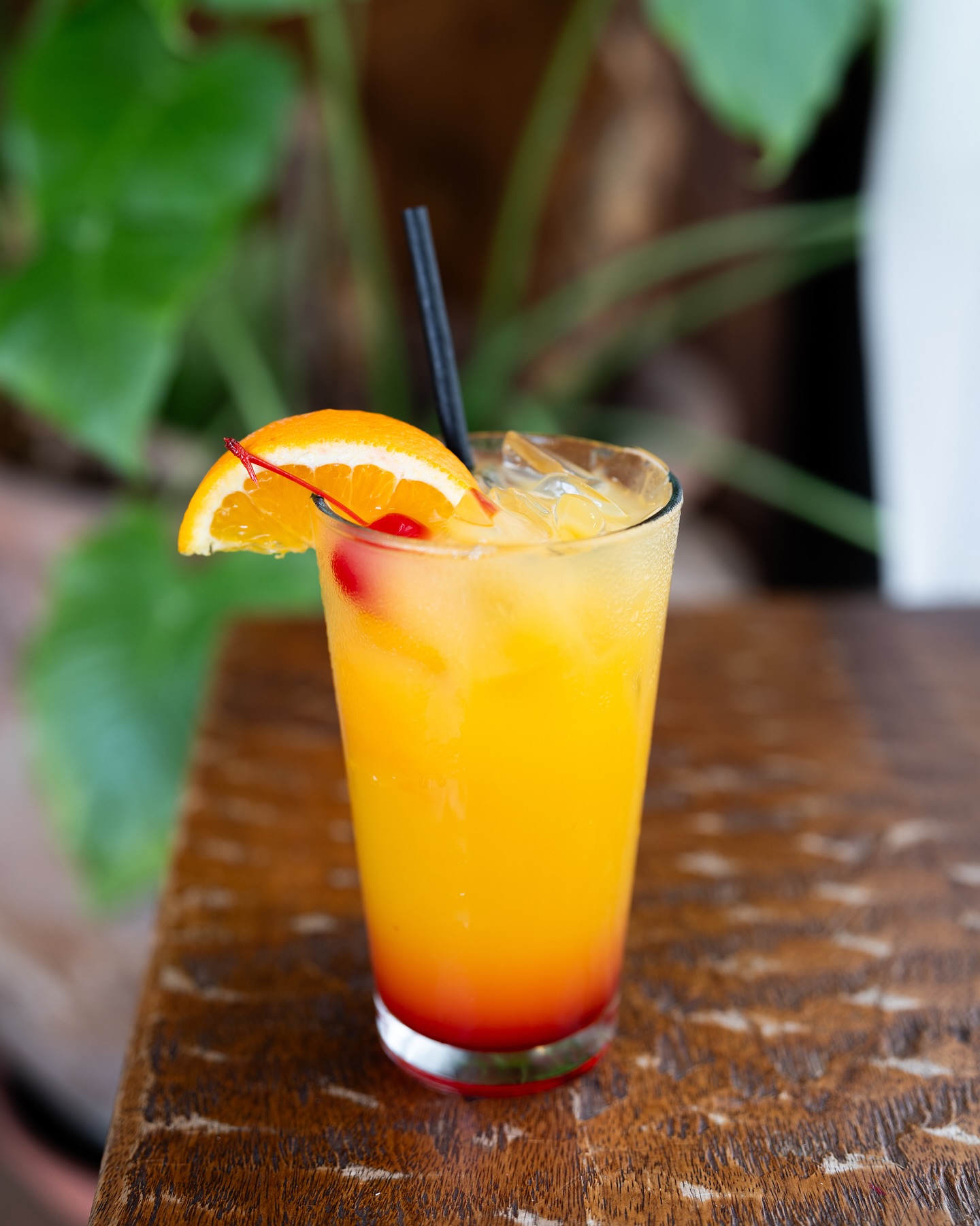 Iced orange and red layered drink with orange slice, cherry, black straw.