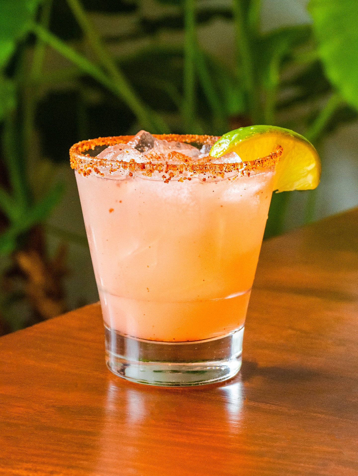 Pink cocktail with spiced rim, ice, and lime wedge on table.
