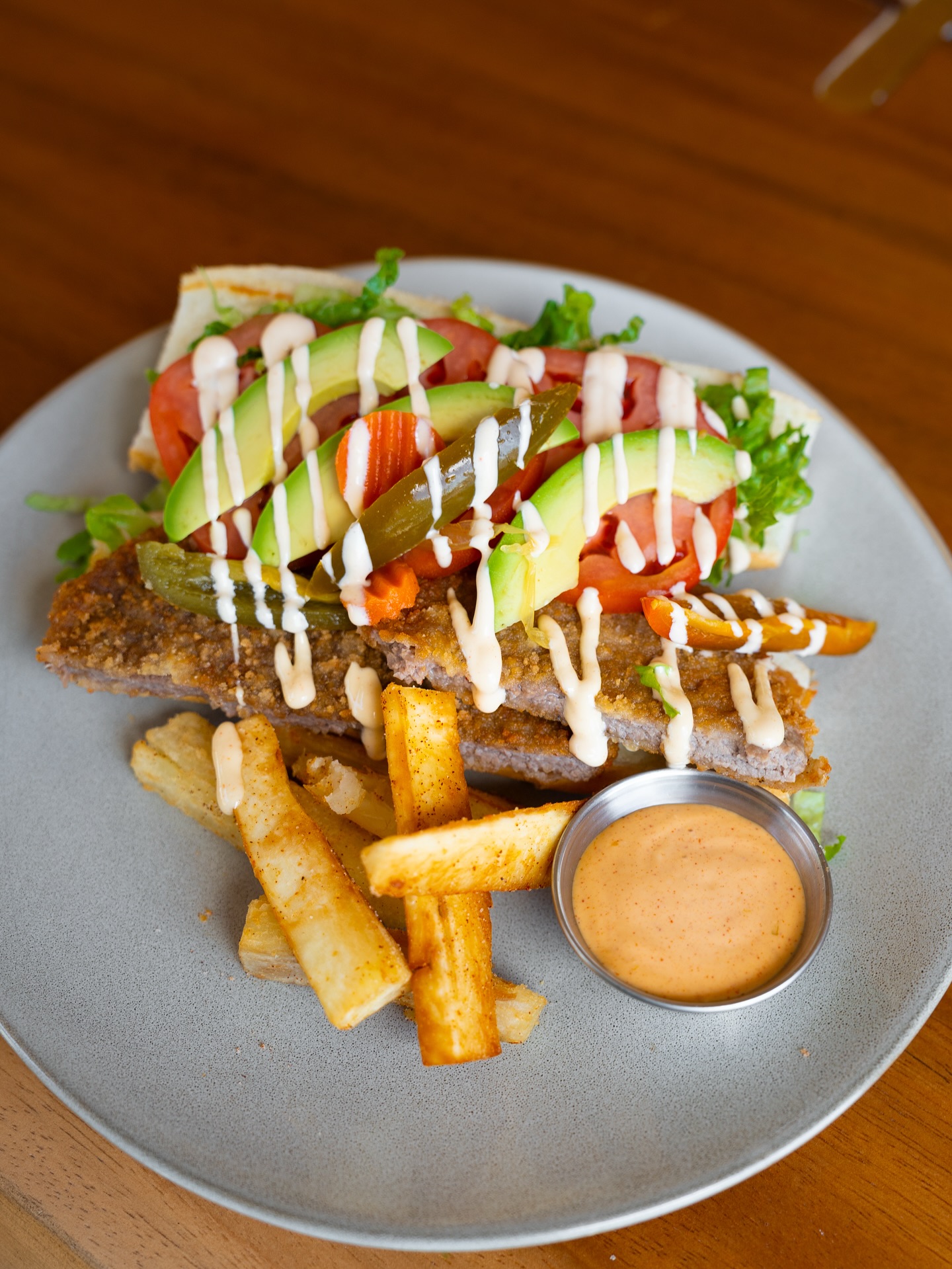 Breaded sandwich with avocado, tomato, pickles, fries, and dipping sauce.
