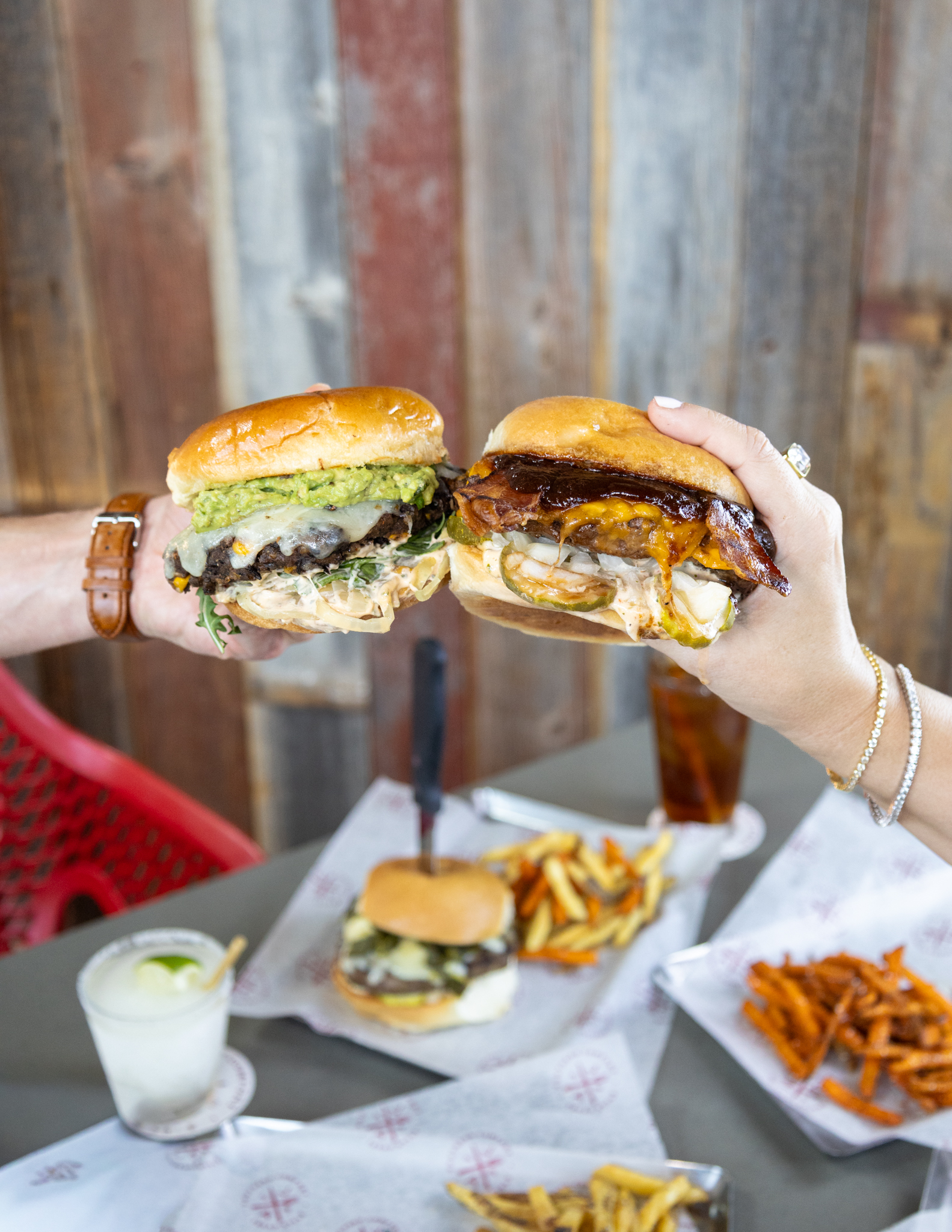 Two hands clinking two loaded burgers above a table of food.