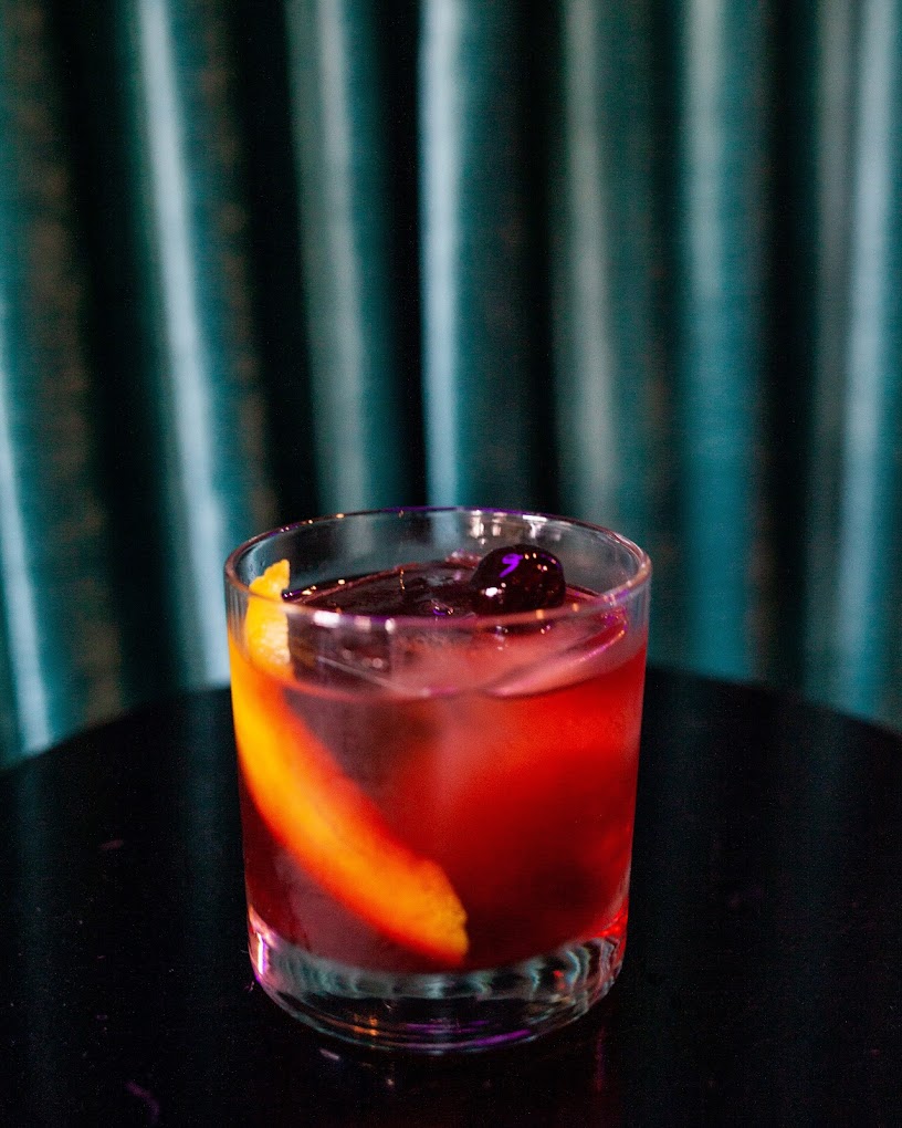 Glass of red cocktail with orange peel and cherry, teal background.