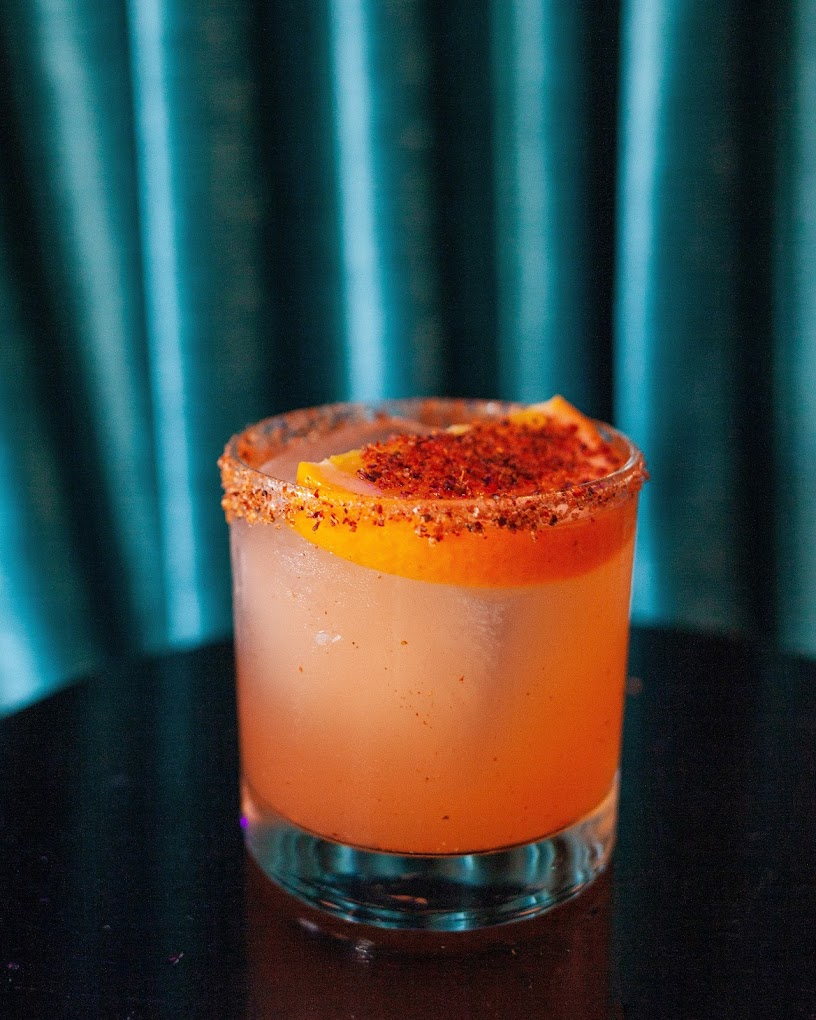 Orange cocktail with spiced rim and orange garnish, teal background.