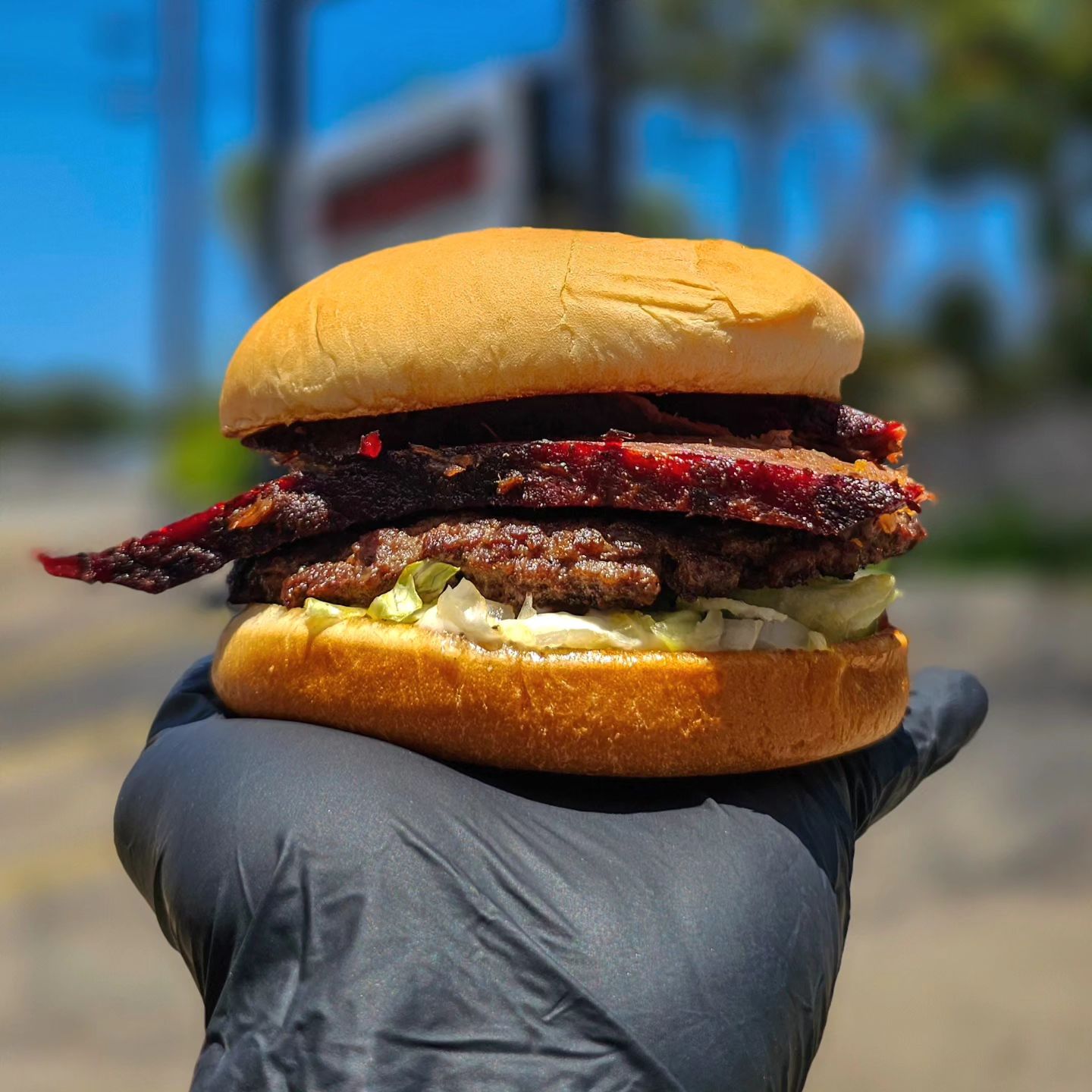 Gloved hand holding a large burger with multiple meats and lettuce.
