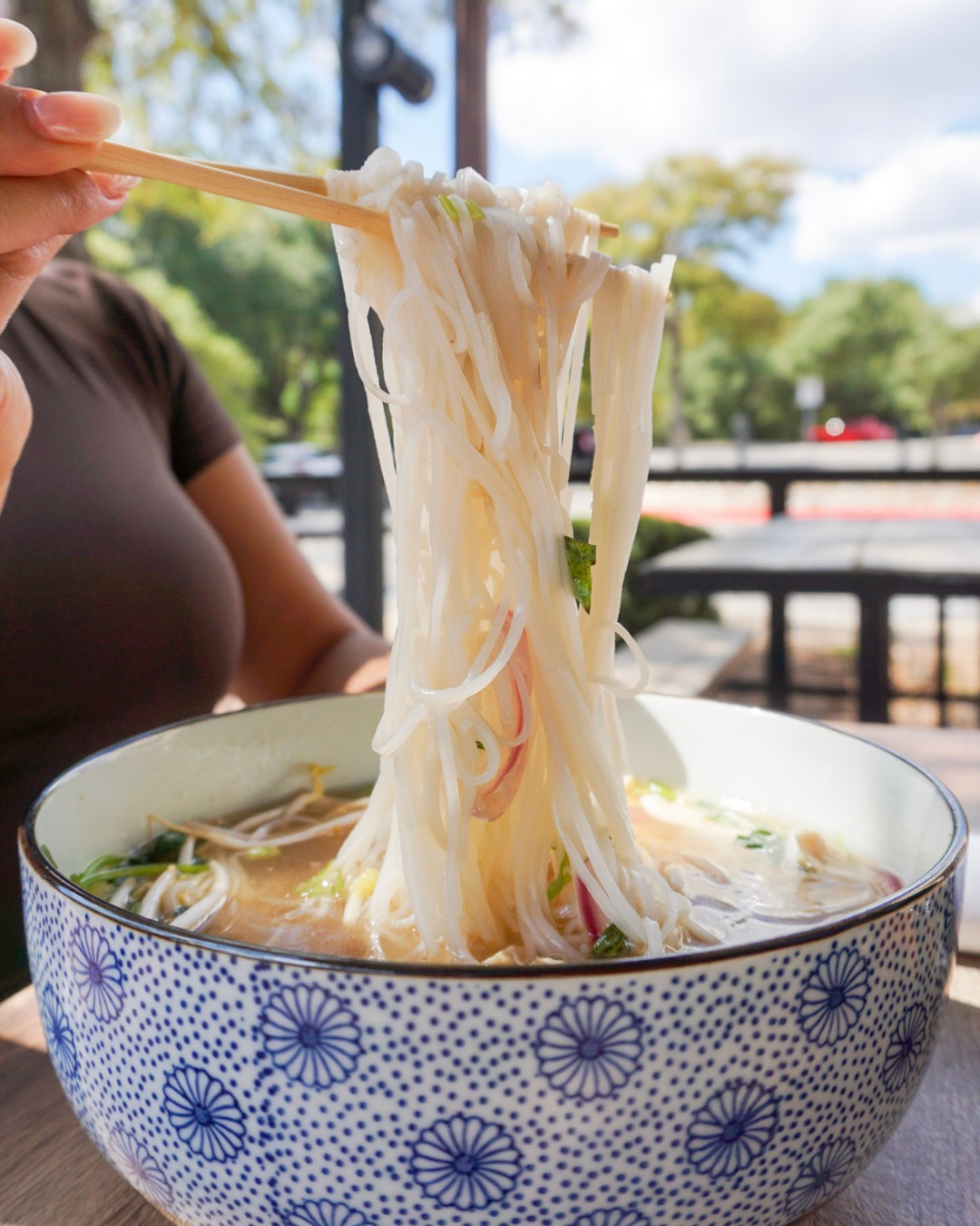 Chopsticks lift white noodles from a patterned bowl outdoors.