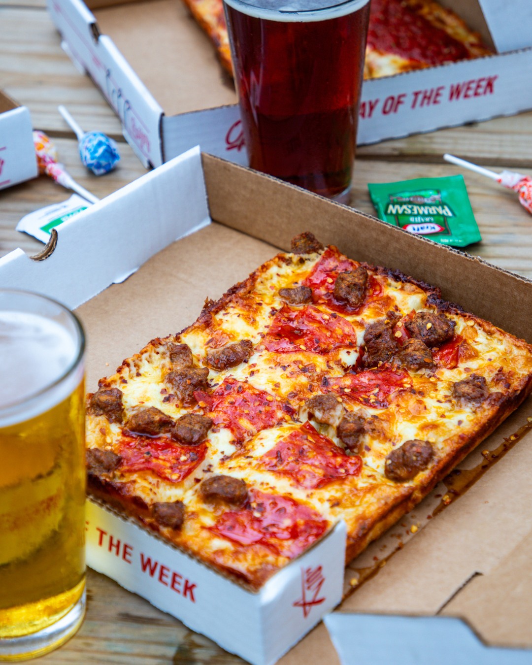 Rectangular pizza with pepperoni, sausage, and two beers on a table.
