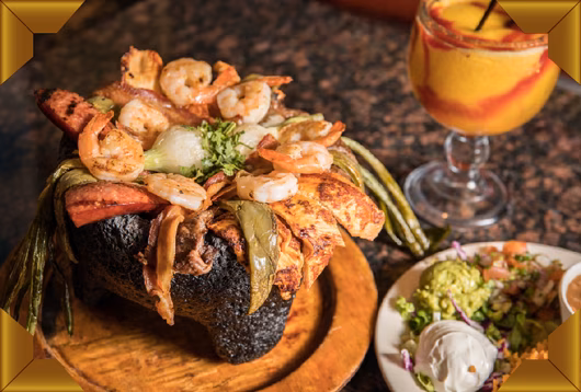 Molcajete meal with shrimp, meat, vegetables, guacamole, and a drink.
