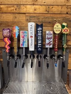 Seven beer taps with assorted brand handles against a wooden wall.