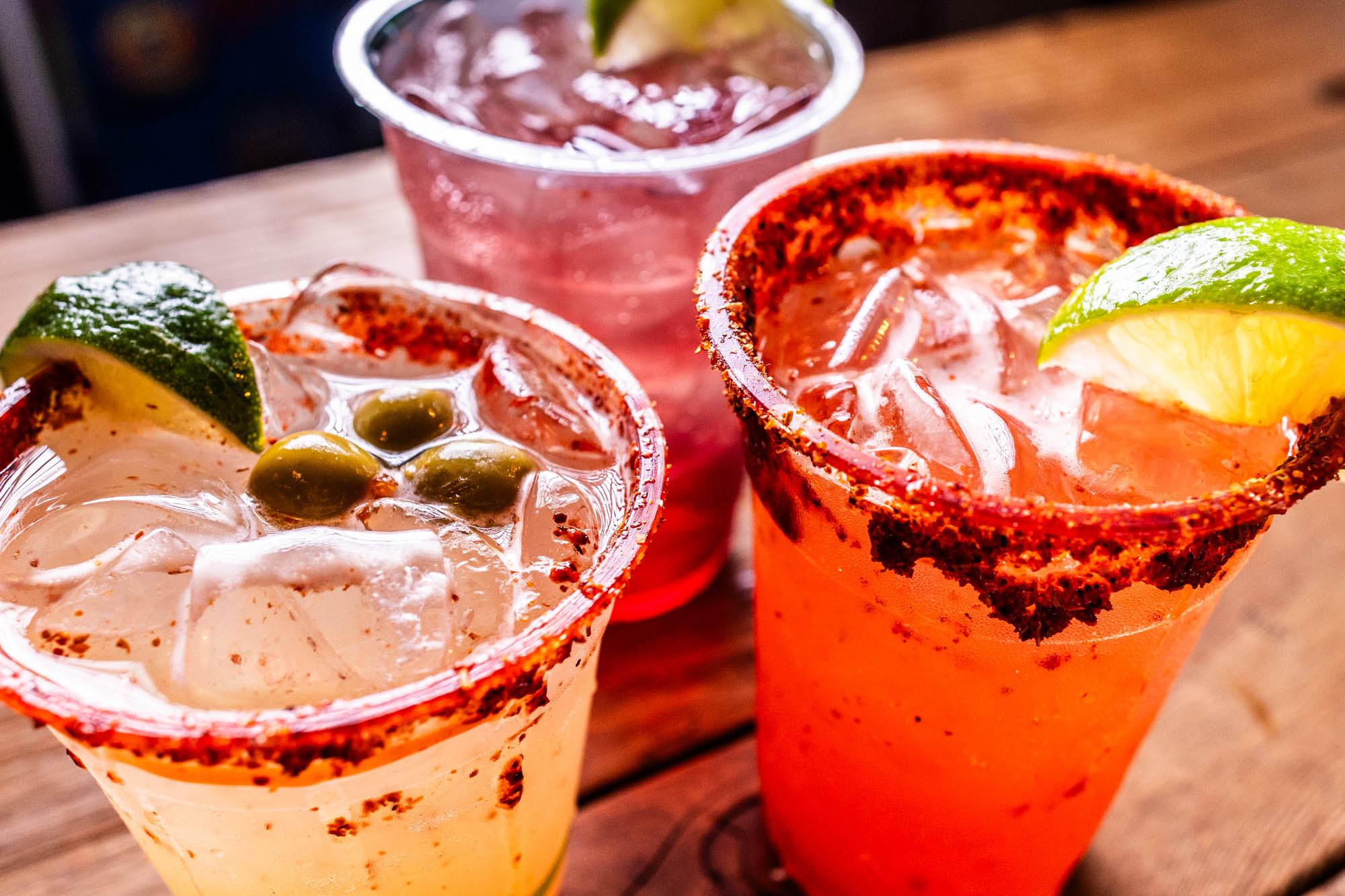 Three colorful iced drinks with lime, olives, and chili-salt rims.