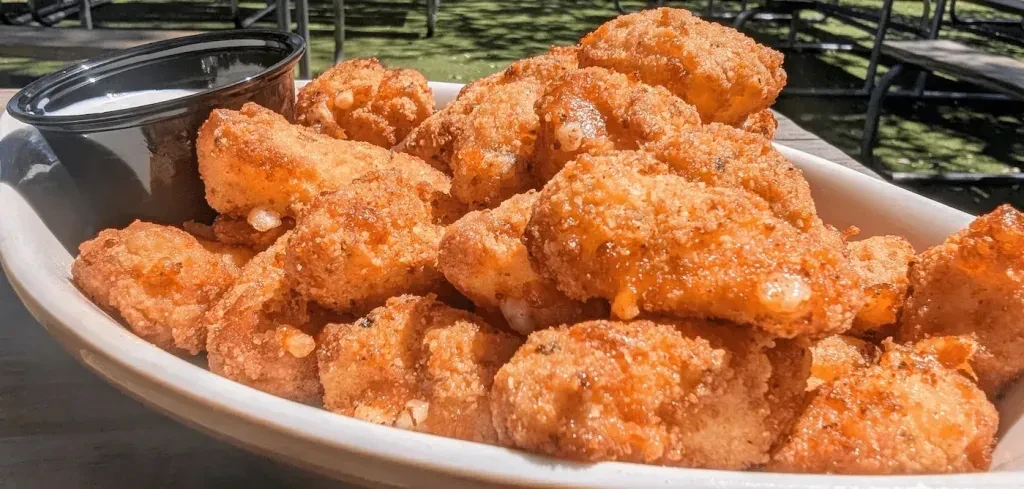 Piled crispy golden-brown fried cheese curds with sauce, in outdoor setting.