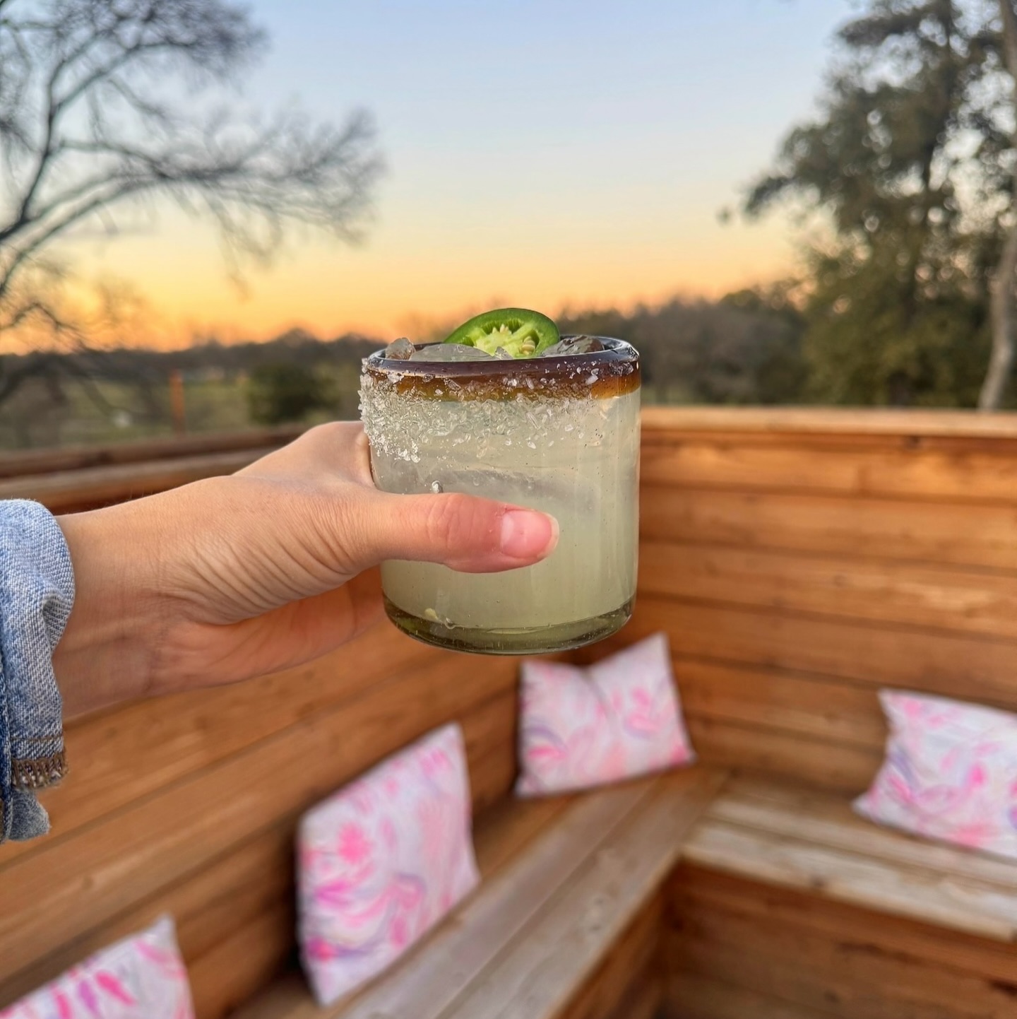 Hand holding a salted cocktail with jalapeño slice, sunset background.