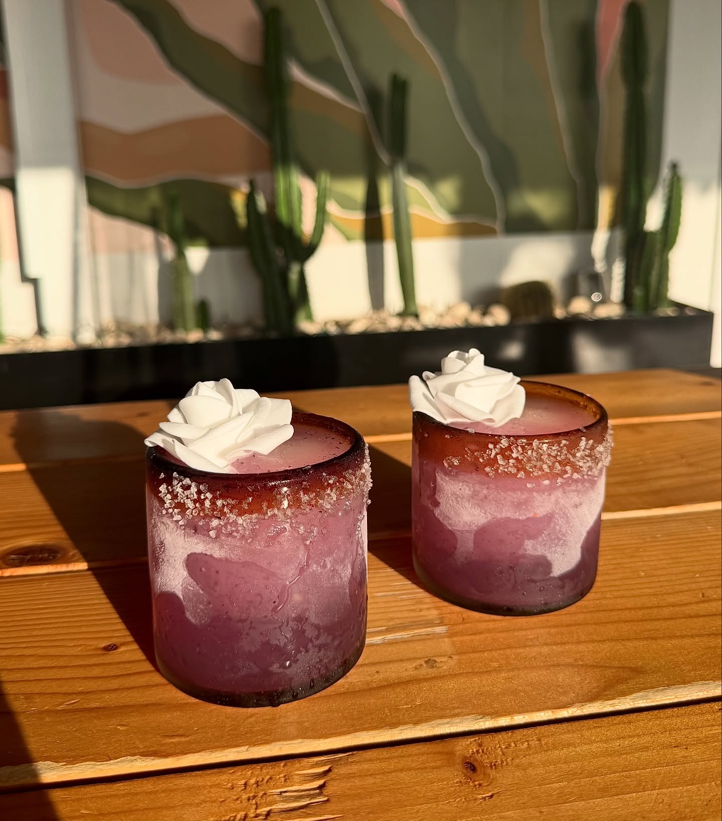 Two purple frosted drinks with rose garnishes on a wooden table.